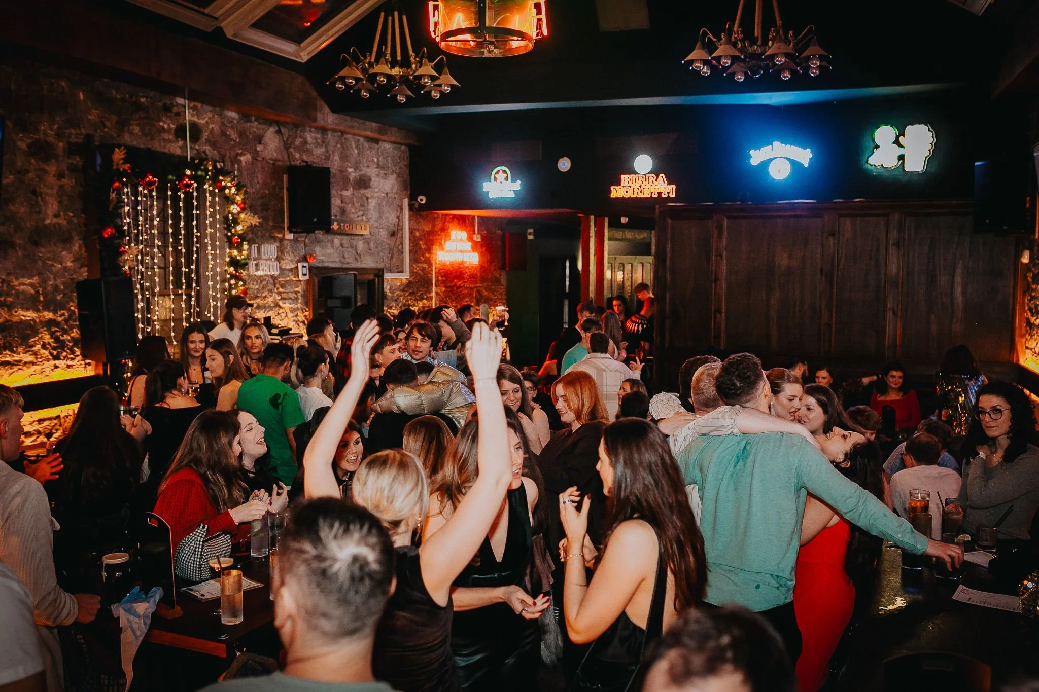 Crowd of people at a lively indoor party or nightclub, dancing and socializing, with warm lighting, neon signs, and festive decorations.