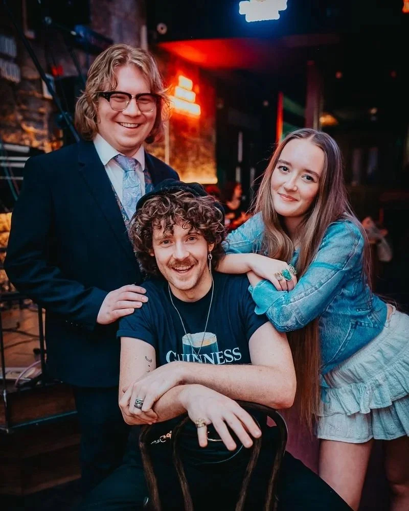 Three young adults posing together in a dimly lit bar or pub, smiling and enjoying their time. One person with curly hair is seated, wearing a Guinness T-shirt, while a woman with long straight hair leaning on his shoulder, and a man with glasses and longer hair standing behind them. The background features neon lights and a relaxed social atmosphere.