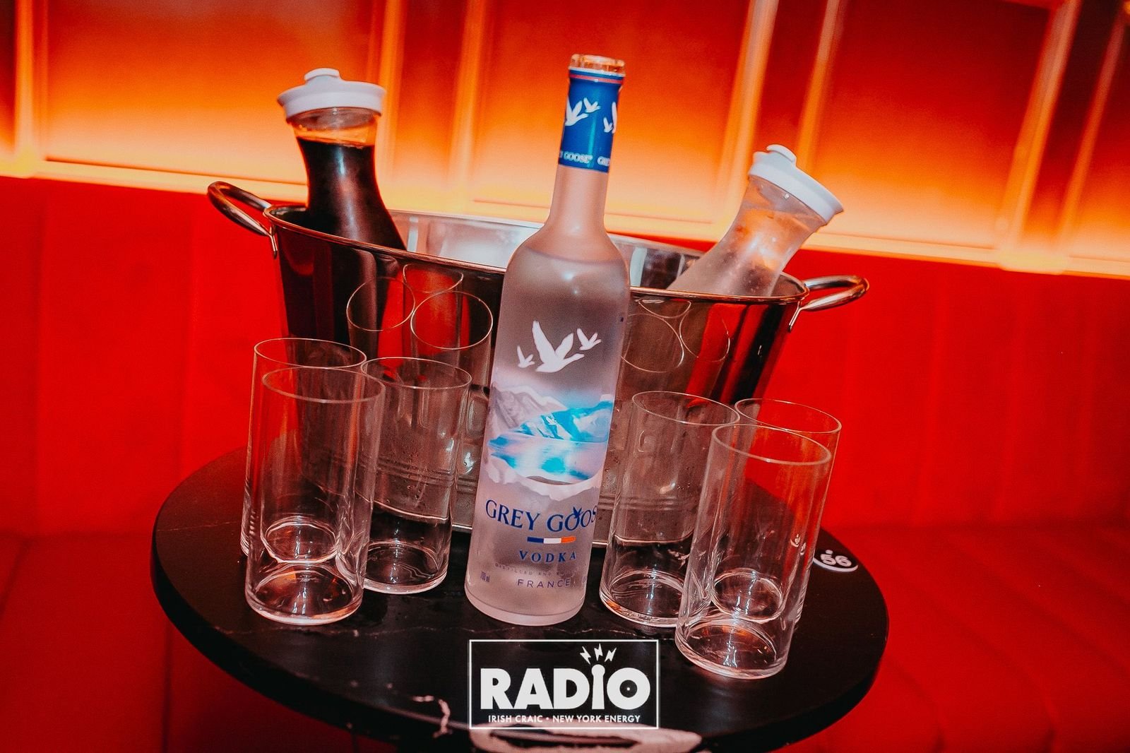 A bottle of Grey Goose vodka surrounded by empty shot glasses, in a metal ice bucket, on a round black table with a 'Radio' logo, against a red background with warm lighting.