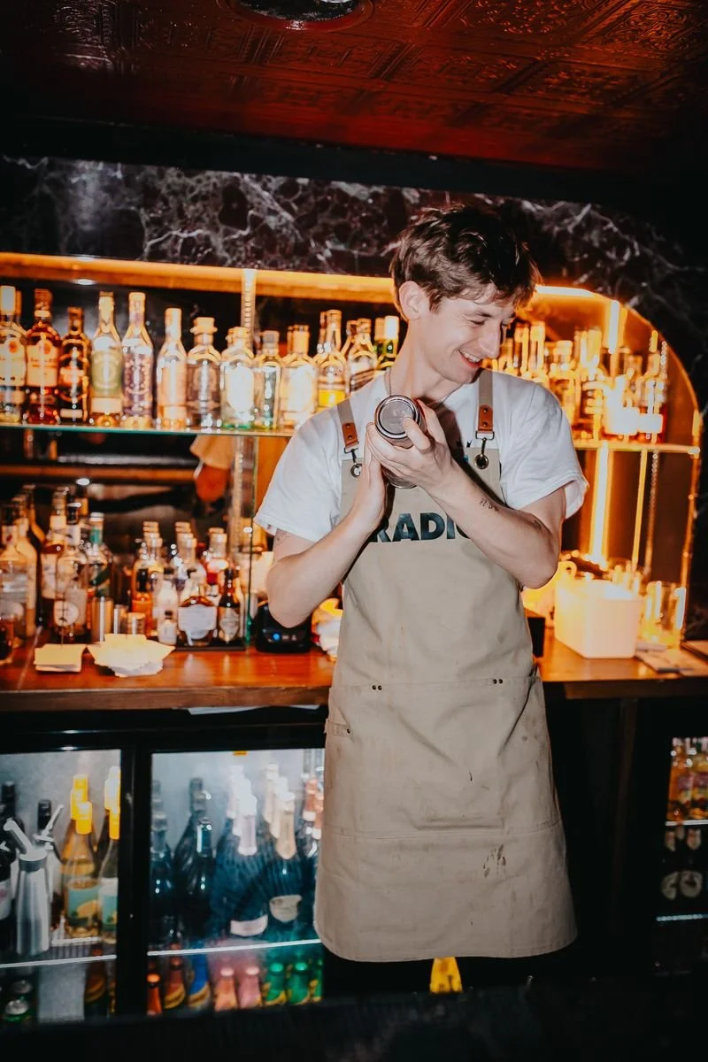 A smiling bartender standing behind a bar with a variety of liquor bottles, wearing a beige apron and a white T-shirt, in a dimly lit bar or club.