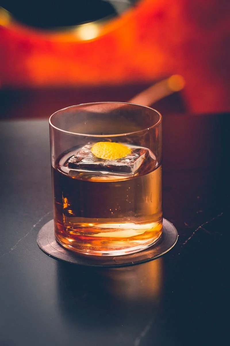 A glass of amber-colored whiskey with an ice cube and a twist of lemon, placed on a dark surface with a blurred warm background.