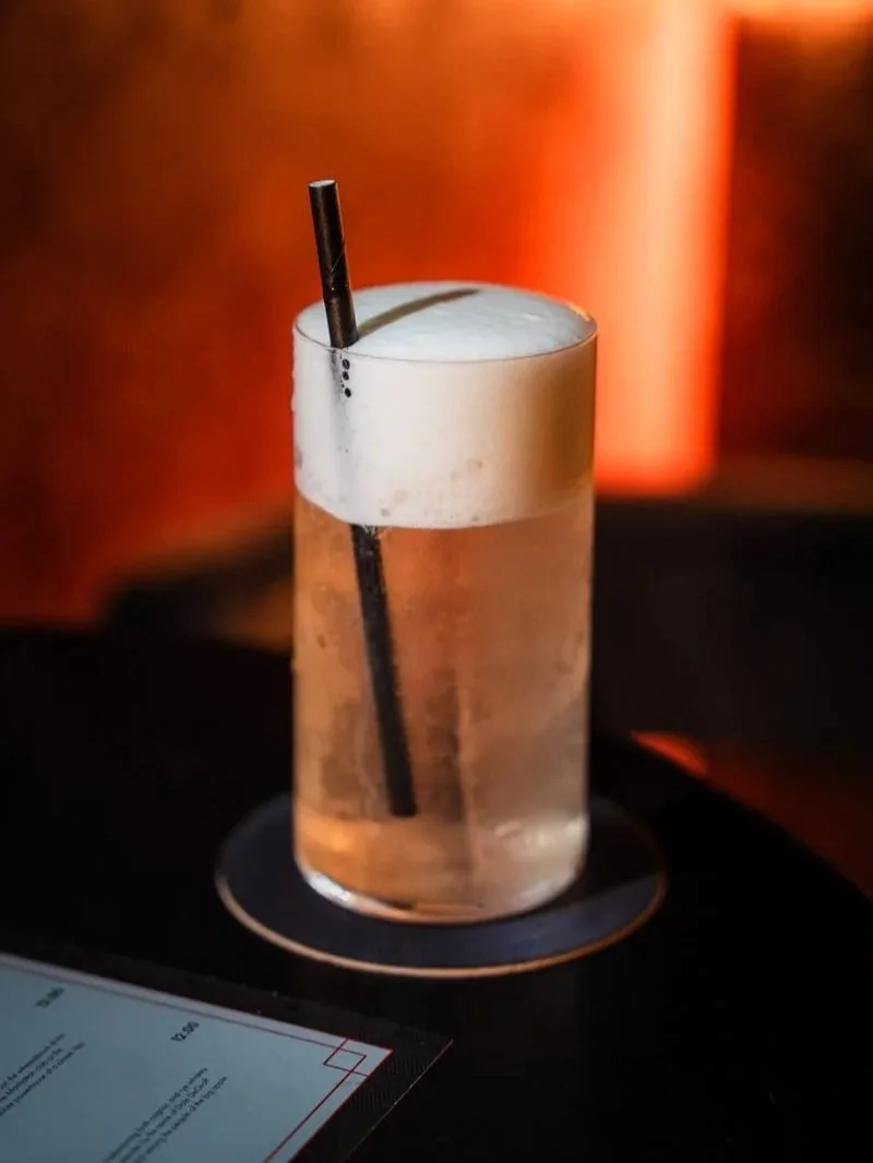 A tall glass of beer with foam on top and a black straw inside, placed on a black coaster on a dark surface, with a blurred warm-colored background.