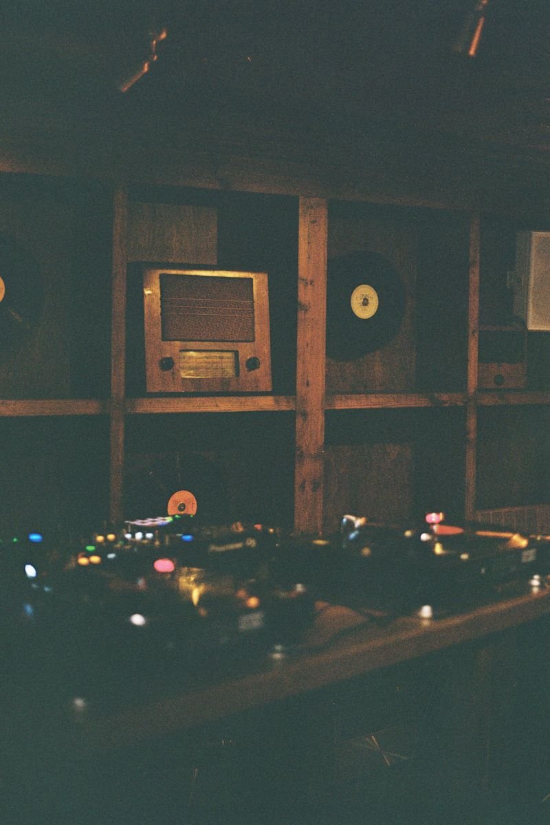 A dimly lit room with a DJ mixer and turntables in the foreground, a vintage wooden radio and a vinyl record on display on the wooden wall in the background.