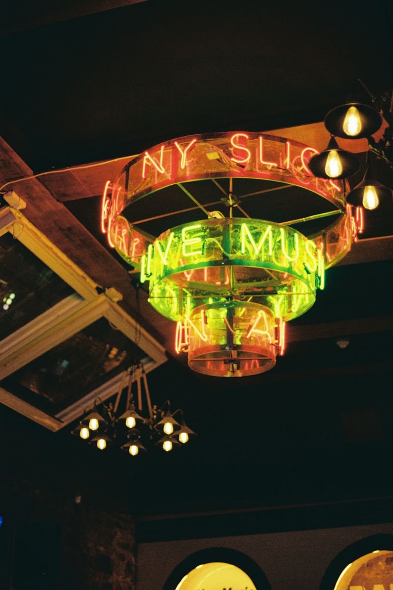 Neon sign in a bar or restaurant with words about NYC, live music, and drinks, illuminated in pink and green colors hanging from the ceiling.