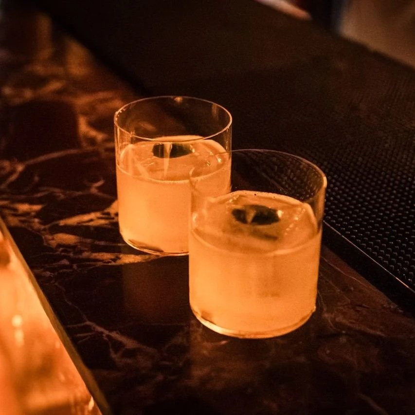 Two short glasses with a light-colored liquid and lime wedges on a dark marble bar counter