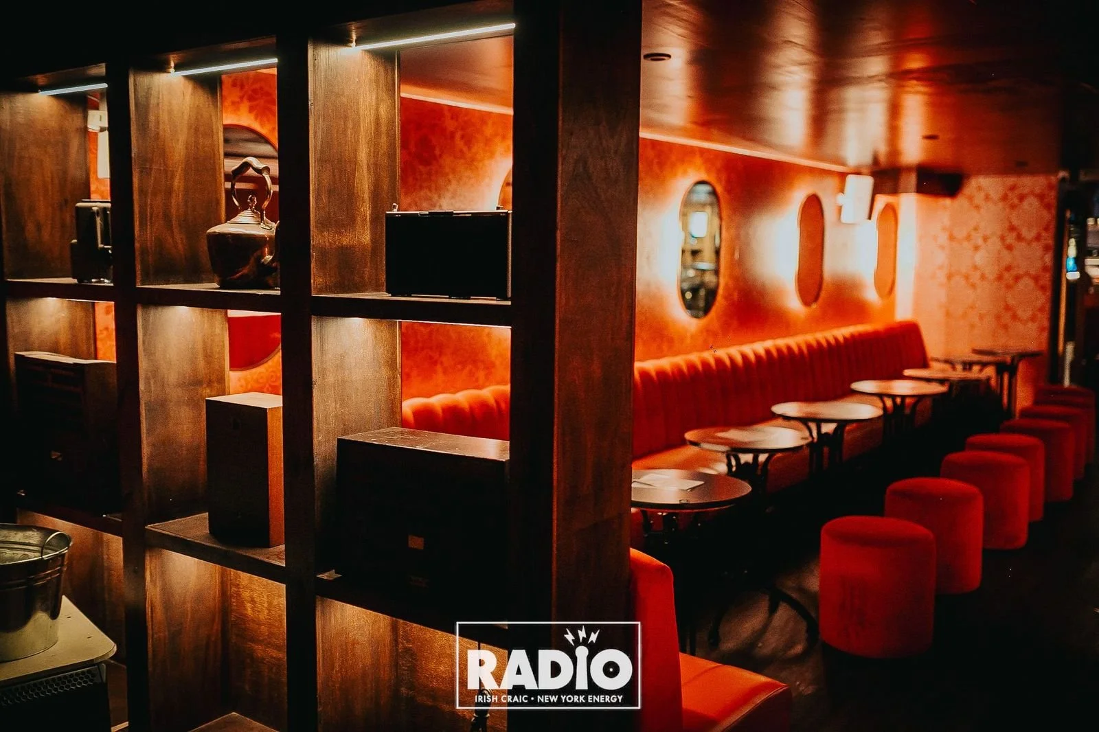 A dimly lit bar or lounge with red and orange tones, featuring a long upholstered red bench along the wall, small round tables, and red stools. Decorative oval mirrors are mounted on the wall.