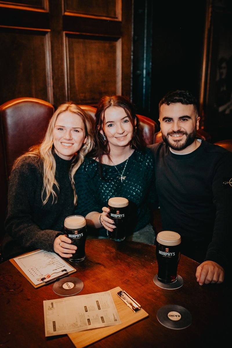 Three friends sitting at a table in a pub, each holding a pint of dark beer, smiling at the camera.