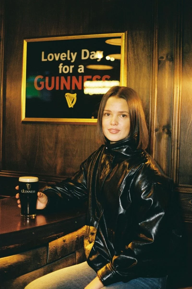 A young woman sitting at a bar holding a pint of Guinness beer, with a sign behind her that reads "Lovely Day for a Guinness", in a pub with wooden paneling.