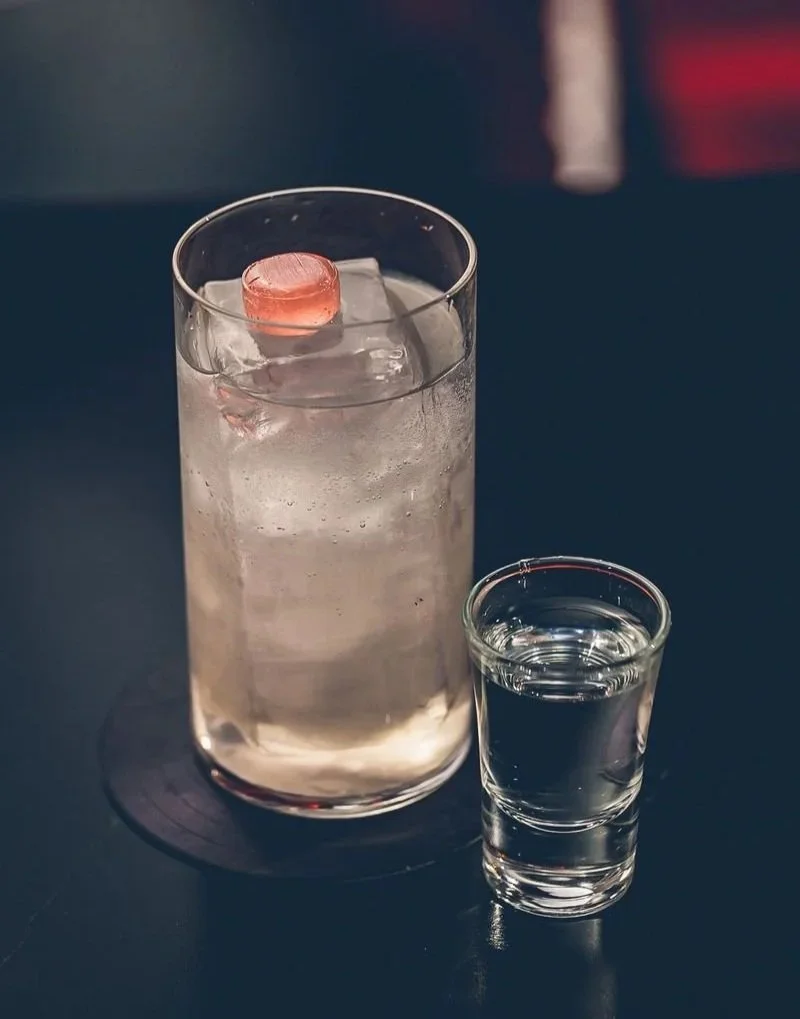 A glass of clear beverage with ice and a pink ice cube, next to a smaller glass of clear liquid, both on a dark surface.