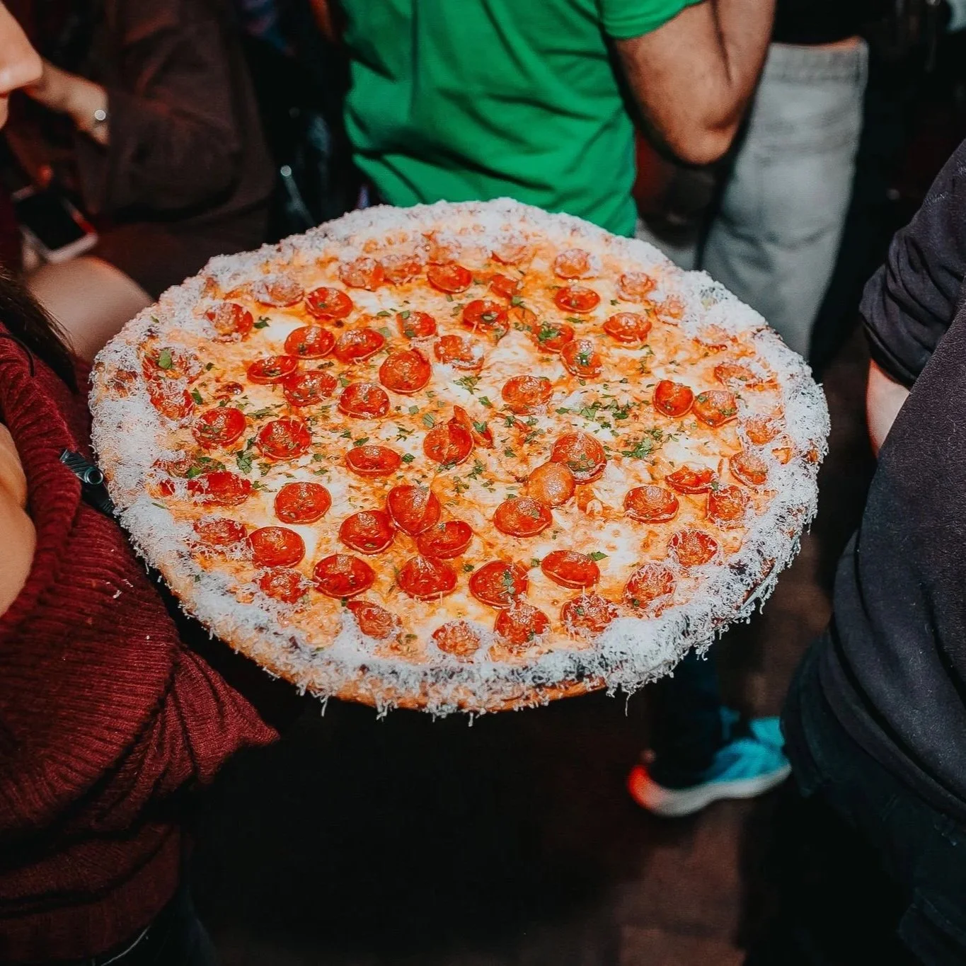 Person holding a large pepperoni pizza with a crust covered in cheese and grated cheese, surrounded by other people at a gathering or party.