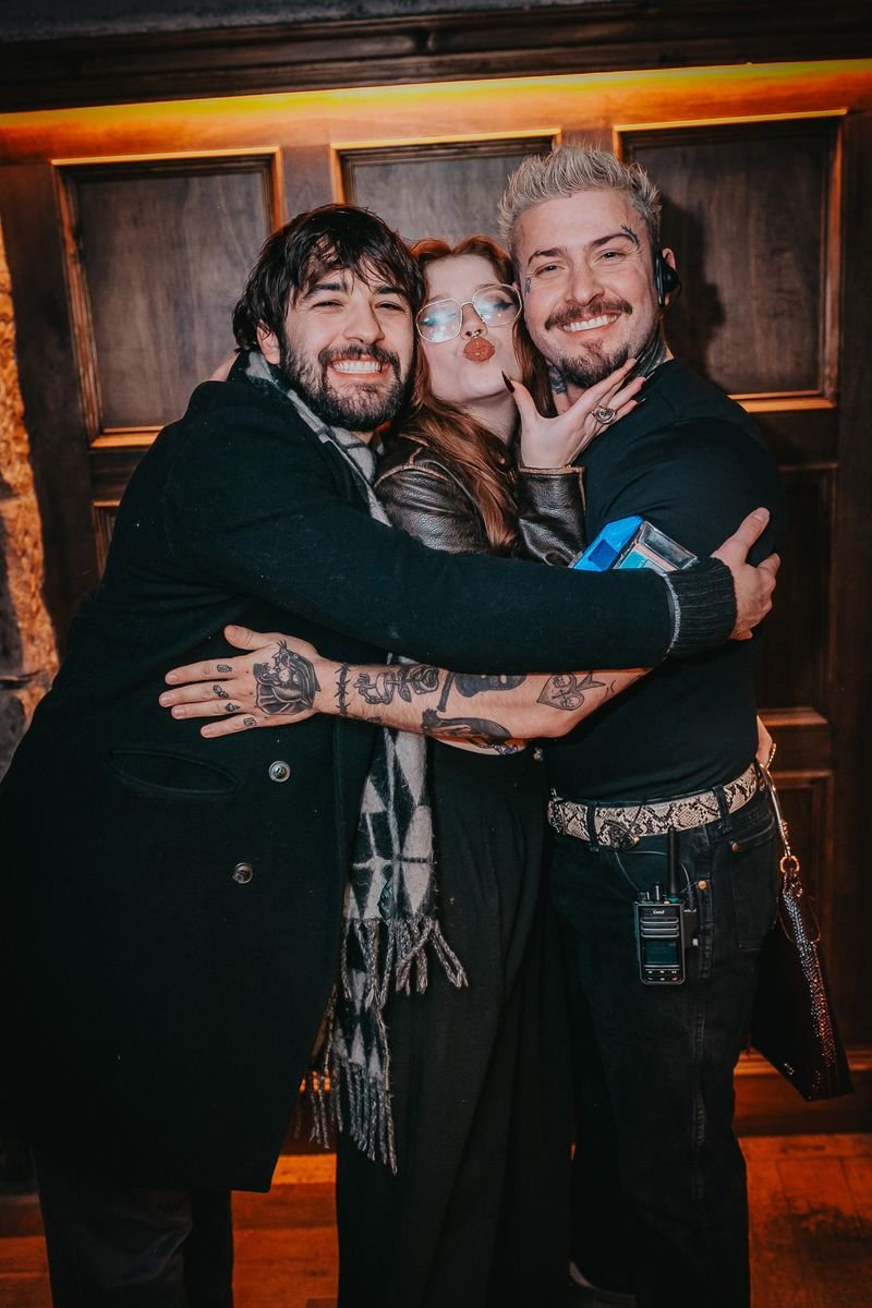 Three friends hugging and posing for a photo, one woman making a kissy face, in a cozy indoor setting with wooden paneling behind them.