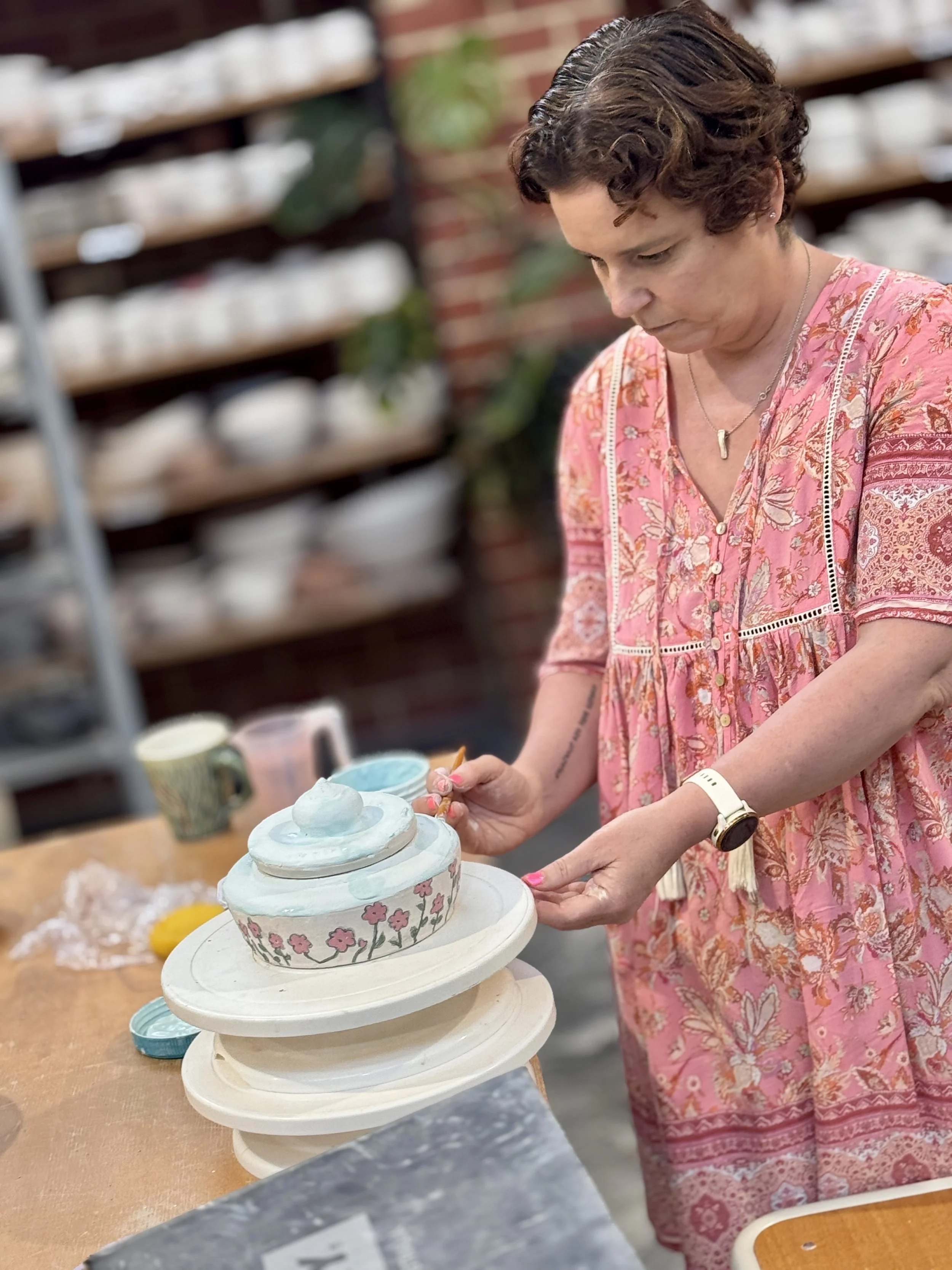 A woman with short curly hair in a pink floral dress decorating a ceramic pot in a pottery studio.