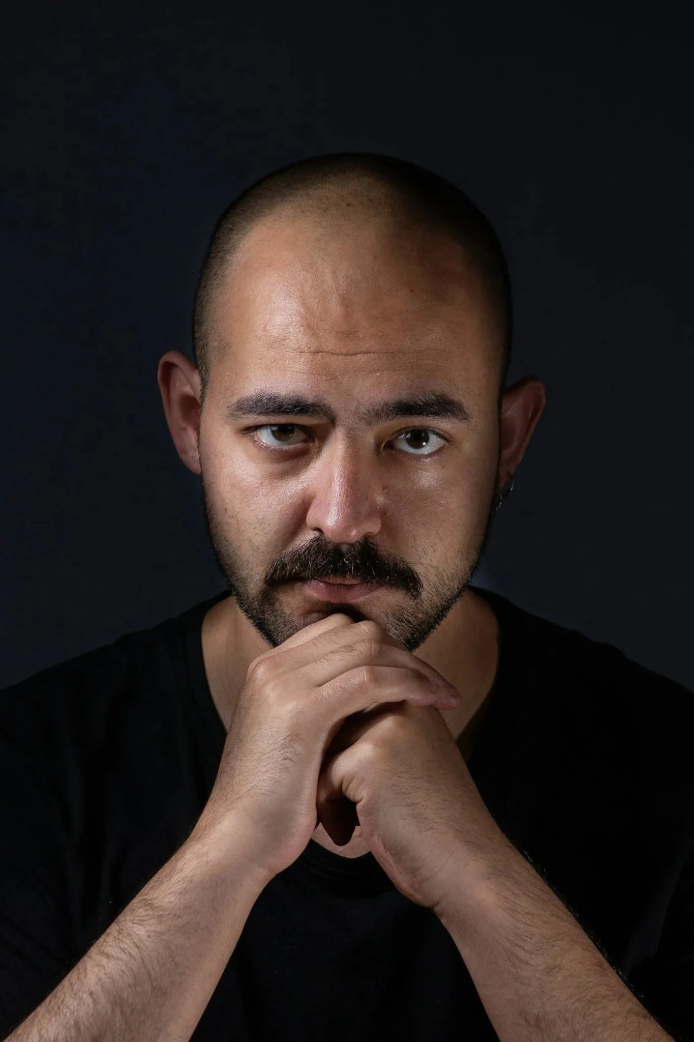 A man with a shaved head, beard, and mustache looks directly at the camera with a serious expression, resting his chin on his hands against a dark background.