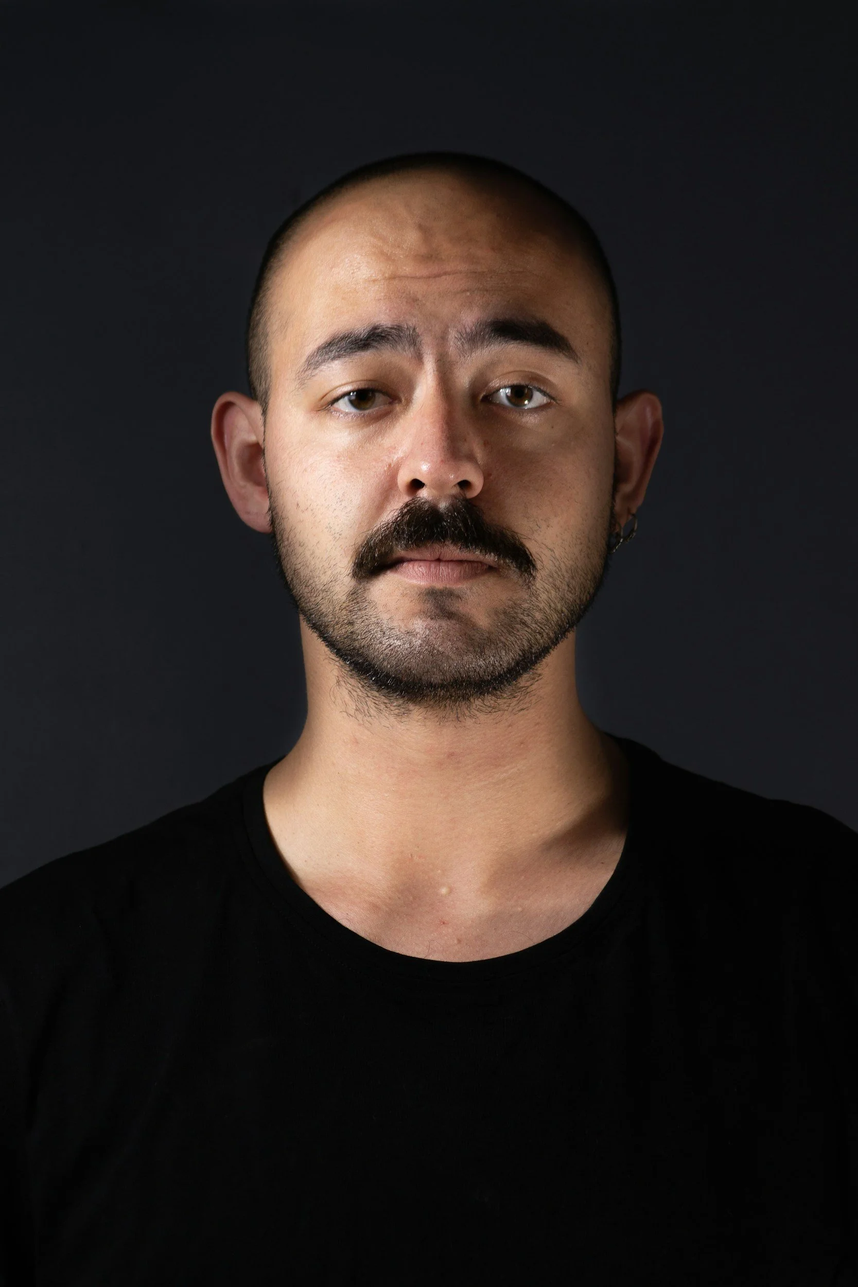A man with a shaved head, mustache, and beard poses against a dark background.