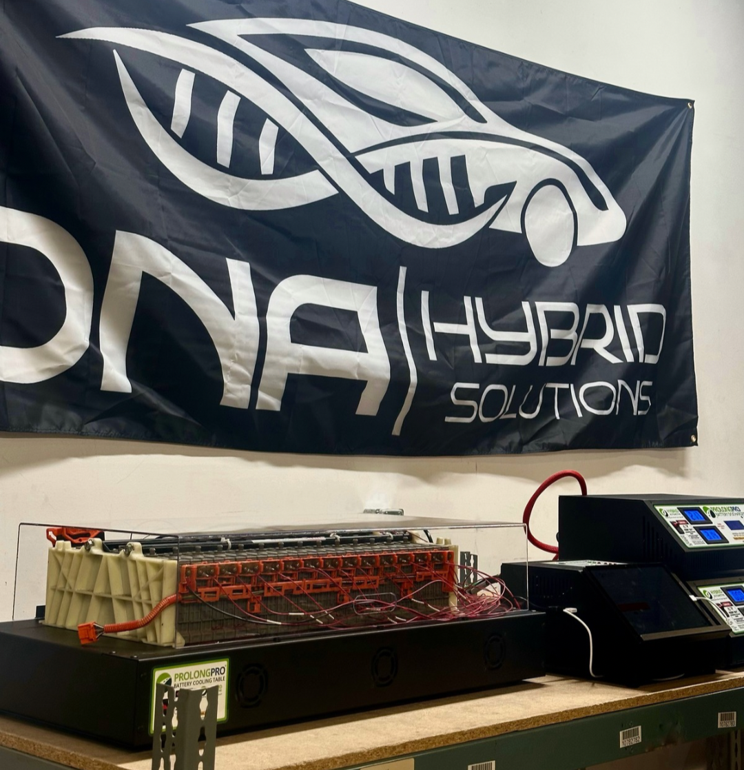 A large flag with a racing car and DNA strand graphic and the text 'HYBRID SOLUTIONS' and 'DYN' hanging on a wall. Below, a testing setup with batteries, wiring, and battery management systems on a workbench.