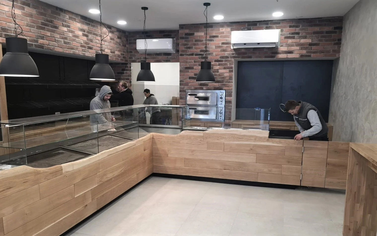 Interior of a modern ice cream shop or cafe under construction, with wooden display counters, black pendant lights, brick walls, and few people working on the setup.