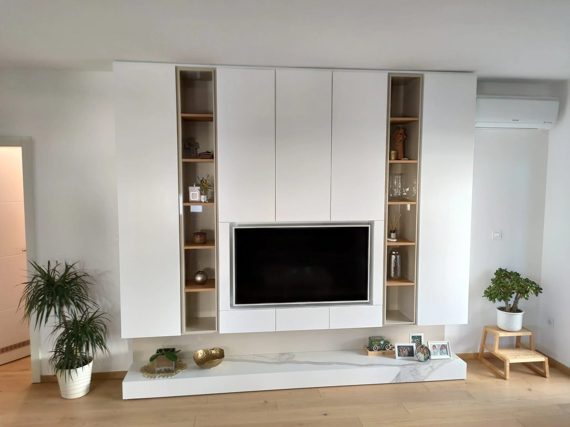 Living room with wall-mounted white entertainment center, flat-screen TV, and decorative items. Potted plants on either side, and a wooden stand with a plant on the right.