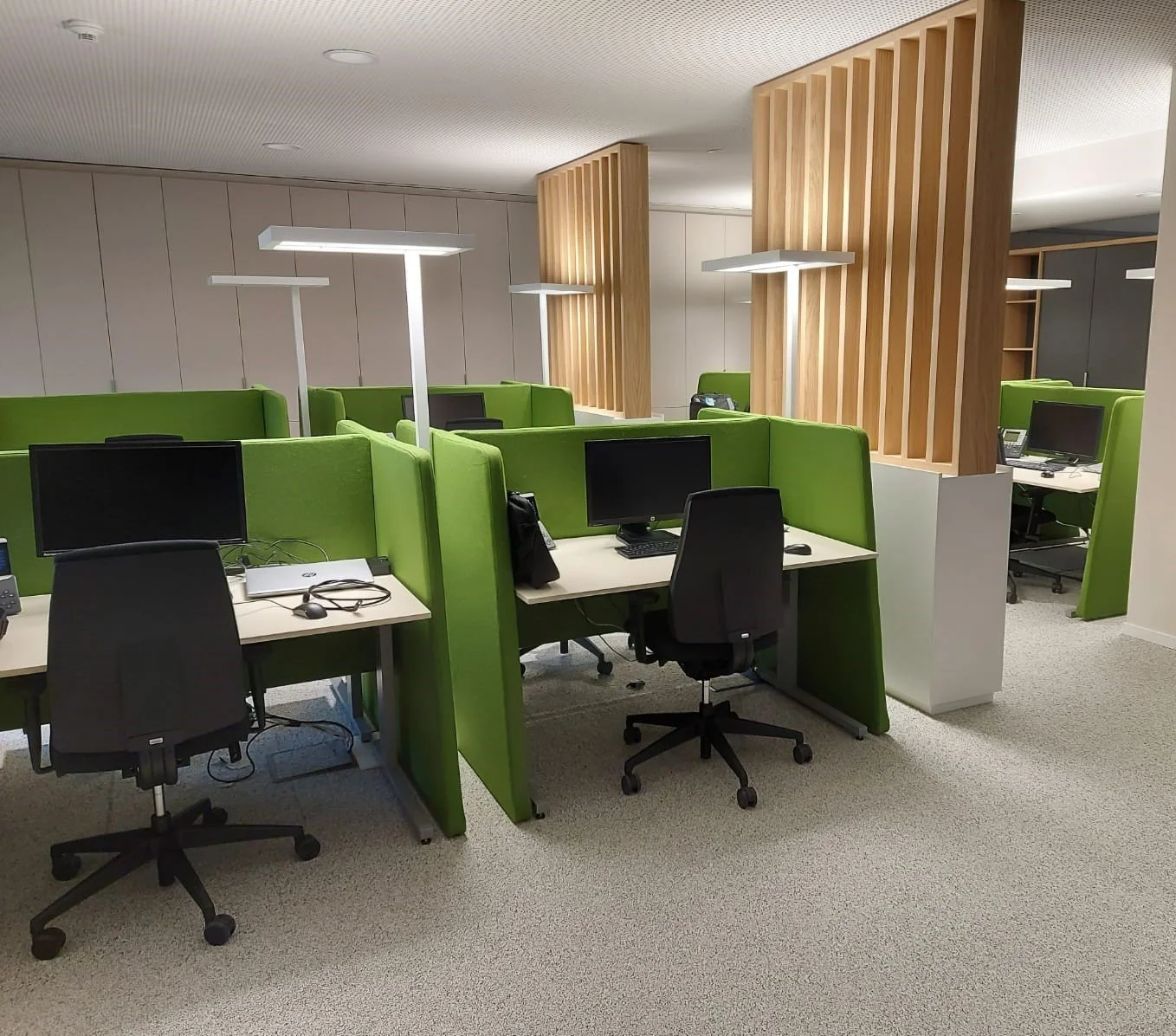 Modern office with green cubicles, black chairs, computer monitors, and white desks. Wooden slat dividers and sleek white lighting fixtures.