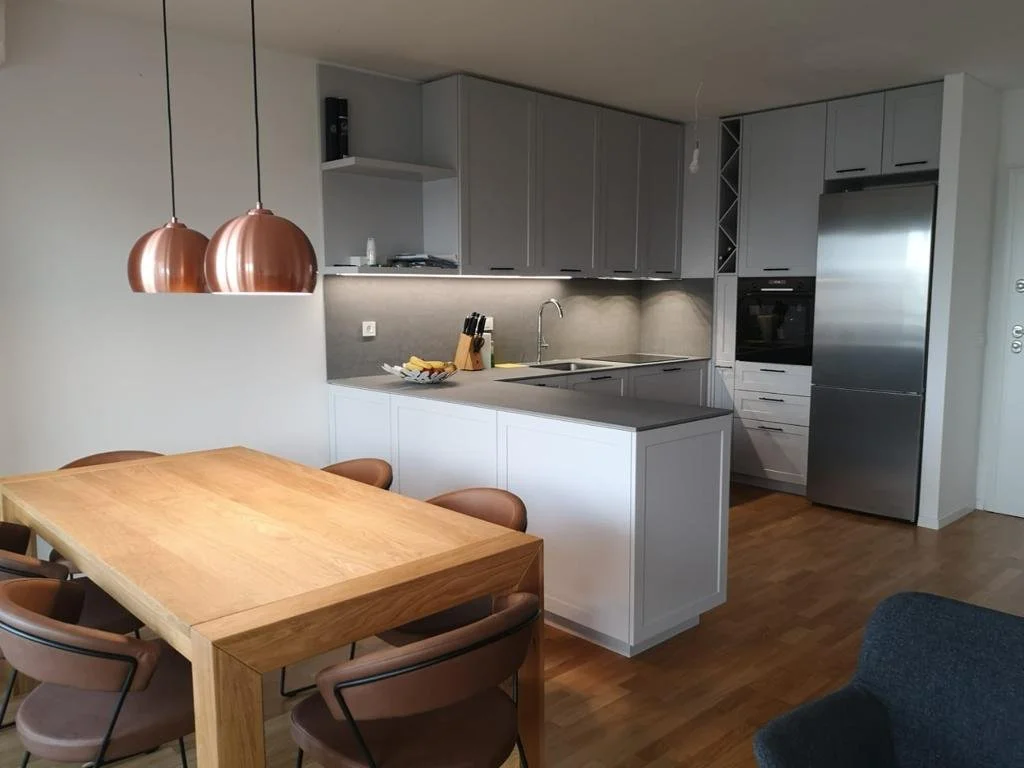 Modern kitchen with gray cabinets, stainless steel appliances including a refrigerator and oven, a kitchen island with a gray countertop, a sink, and a wooden dining table with six brown chairs. Copper pendant lights hang above the dining table.