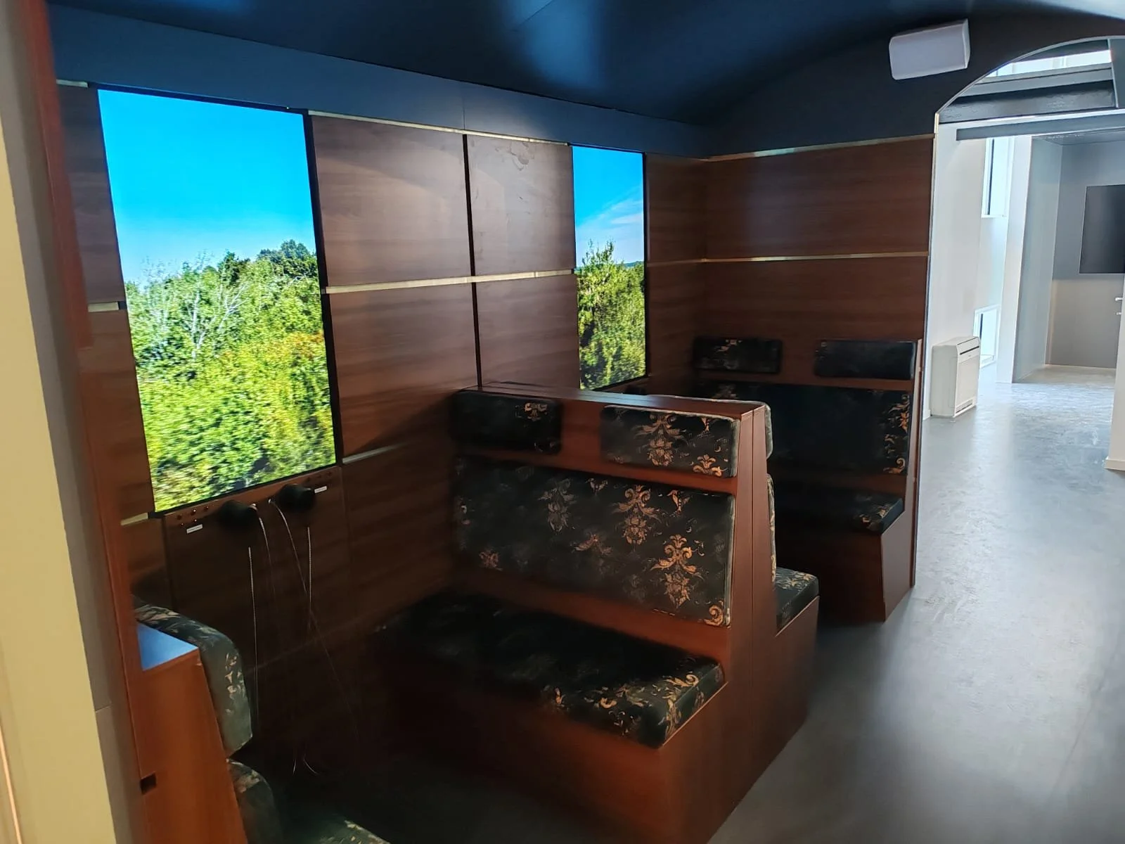 Indoor cafe or lounge with wooden wall panels, large digital screens displaying a nature scene, and floral-patterned upholstered booths.