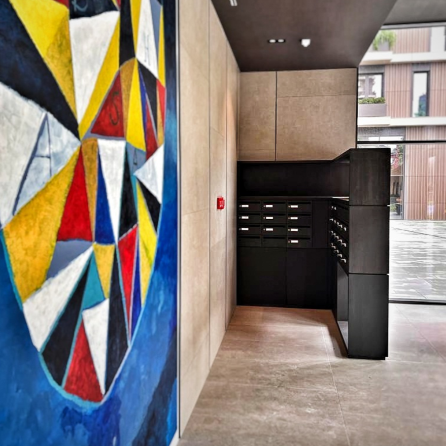 Interior corner of a building with a colorful abstract painting on the left wall, beige tiled walls, a black mailbox and package lockers, and large glass windows revealing outdoor balconies.