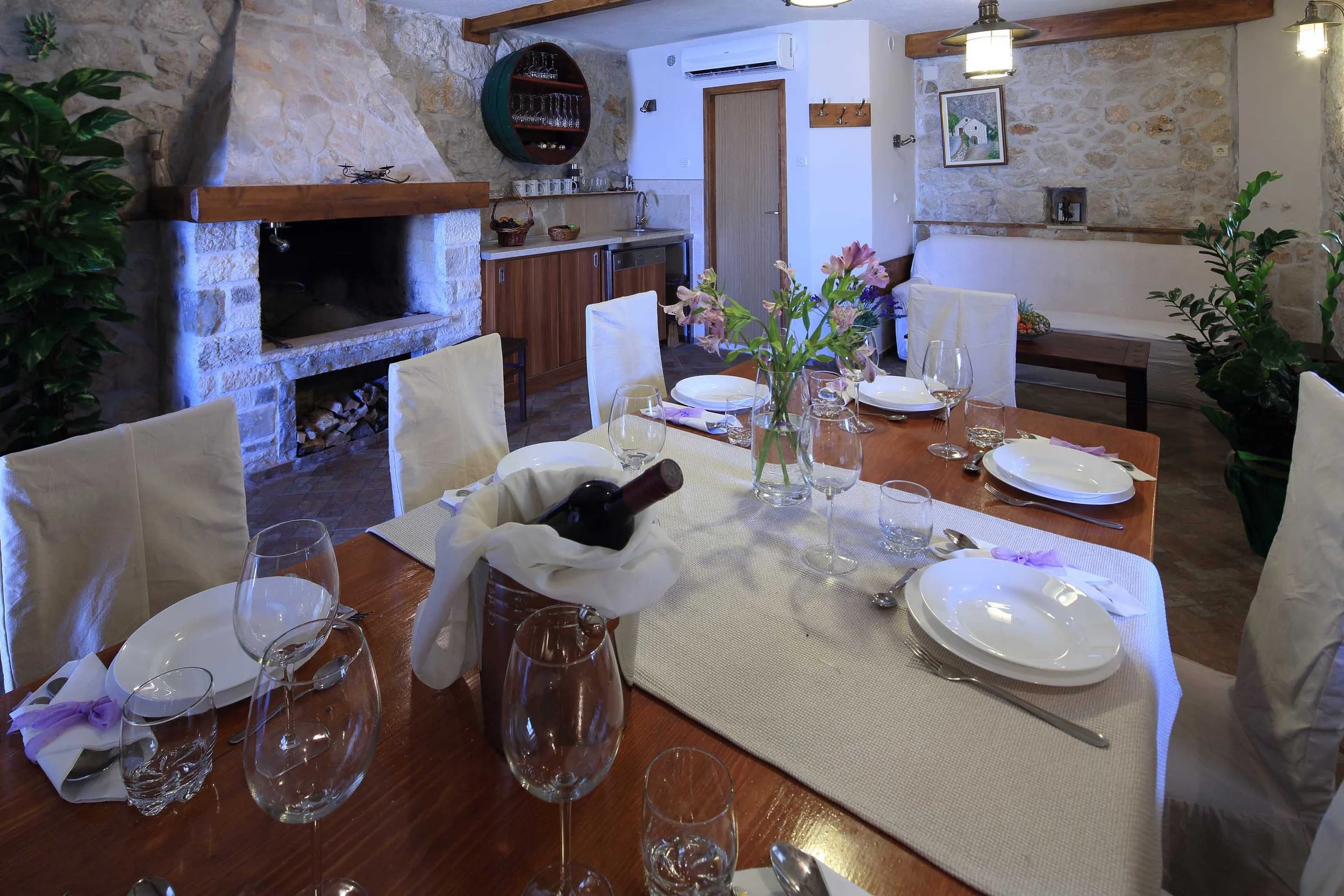 A cozy dining room with a large wooden table set for a meal, decorated with a vase of flowers. Surrounding the table are white-covered chairs. The room features a stone fireplace with a wooden mantle, a built-in wood storage, and a kitchenette area w