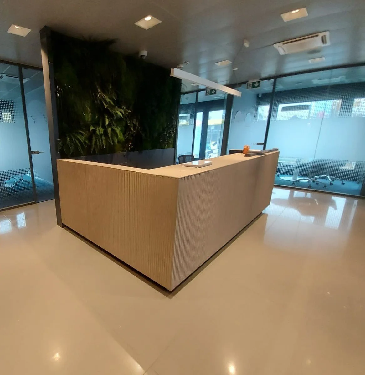 Modern office reception area with a curved wooden desk, a green wall, and glass walls with frosted design, illuminated ceiling lights, and a view of the street outside.
