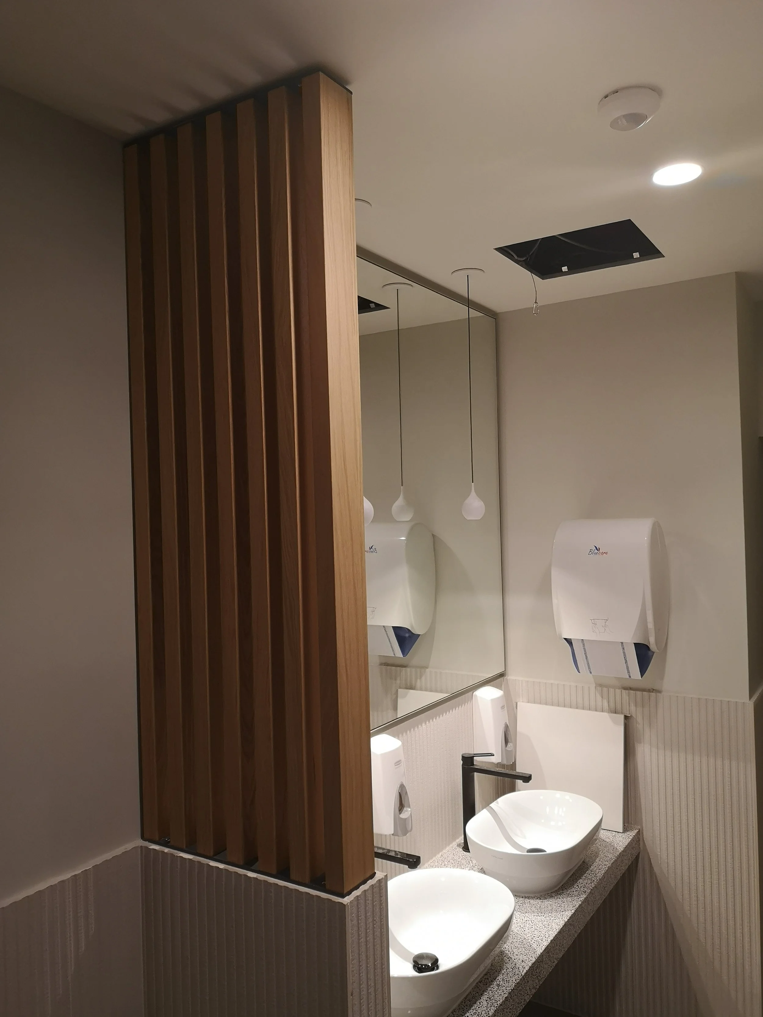 A modern bathroom with a mirror, a white sink with a black faucet, a soap dispenser, a paper towel dispenser, hanging pendant lights, and a wooden slatted partition. The ceiling has recessed lighting and a ventilation grille.