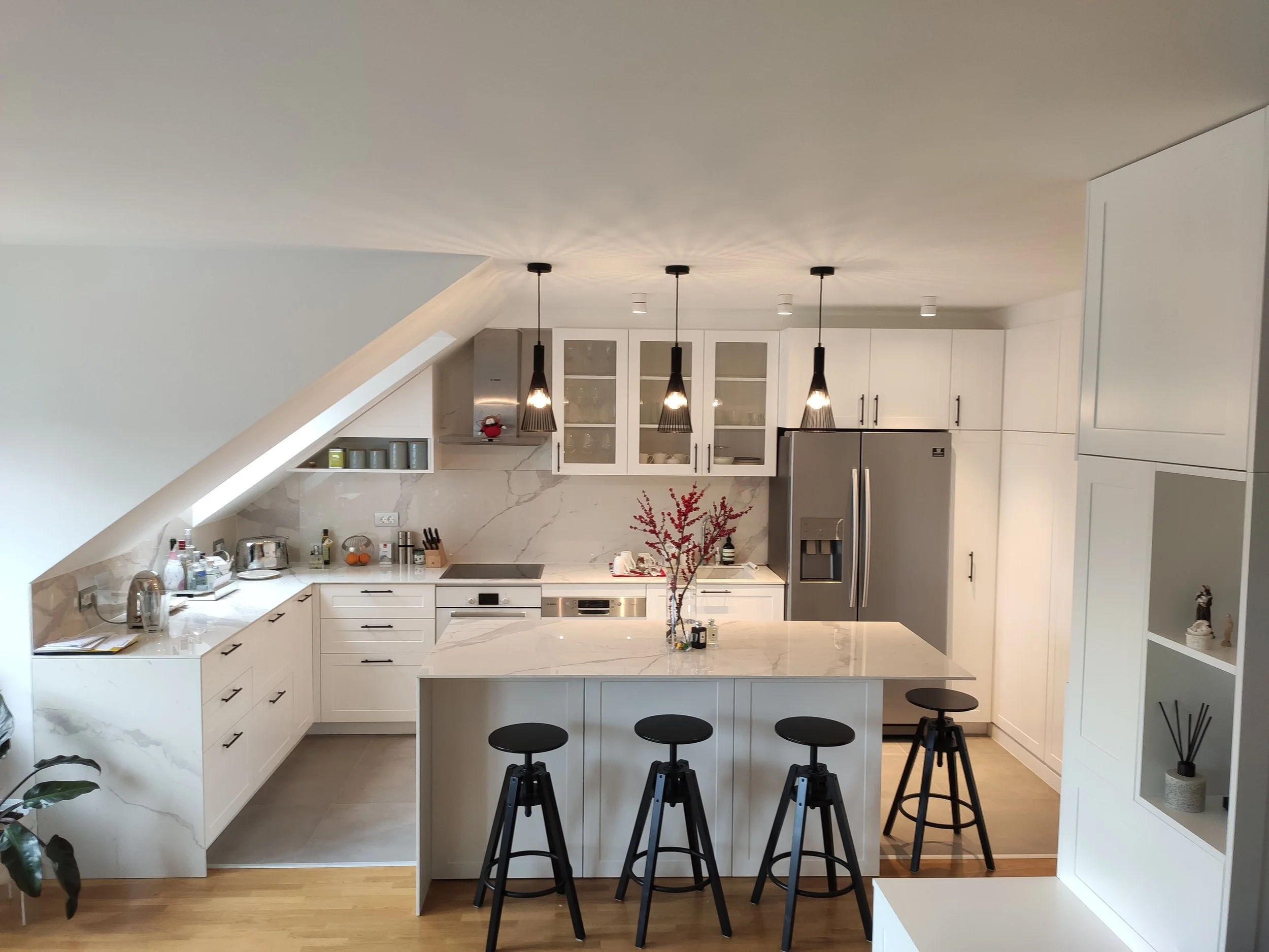 Modern white kitchen with marble countertops, black pendant lights, stainless steel appliances, a central island with chairs, and decorative items on shelves.