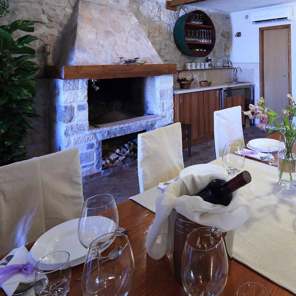 A dining room with a table set with wine glasses, plates, and napkins, featuring a wine bottle in a bucket wrapped with a cloth, with a fireplace, wood logs, and a kitchenette in the background.