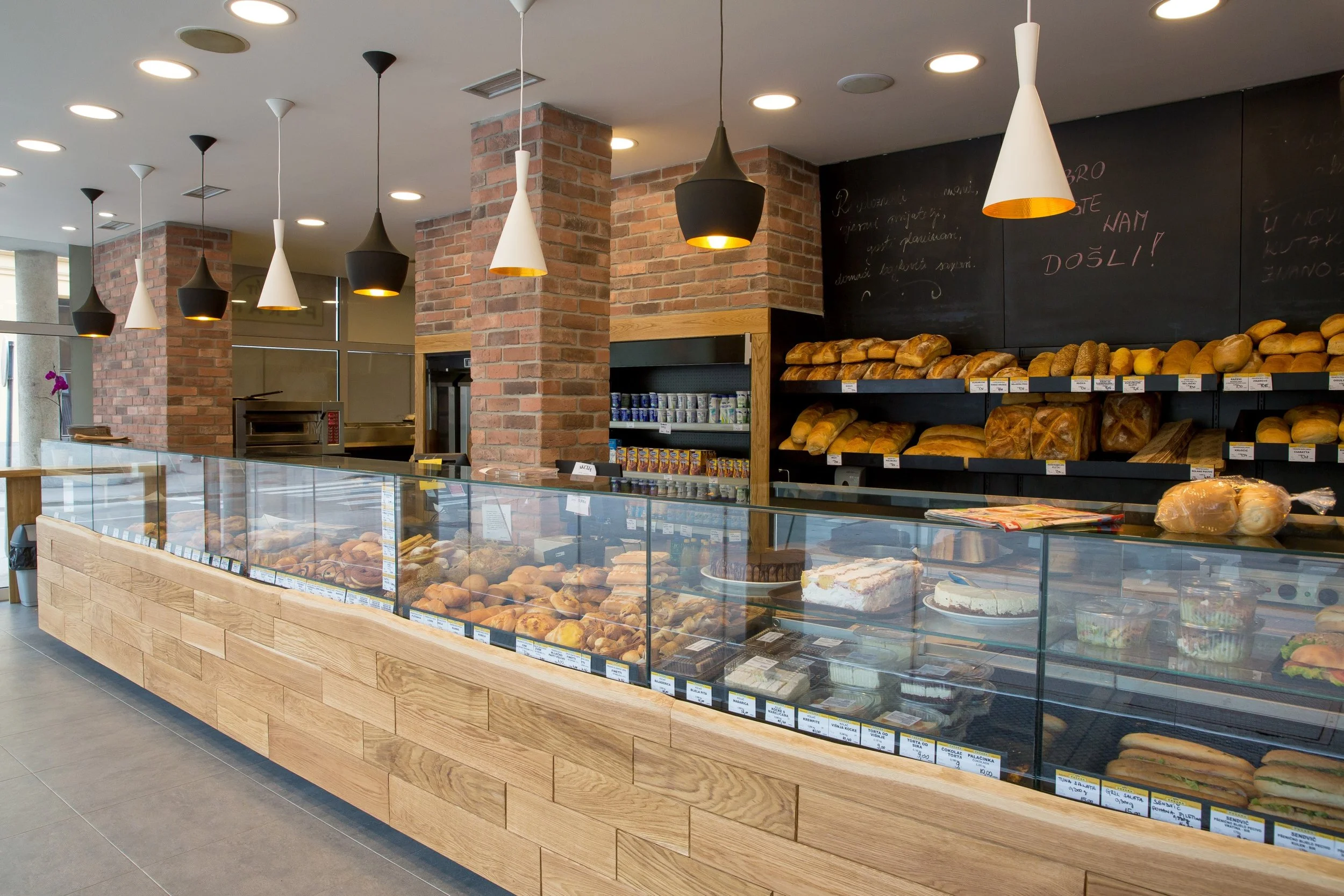 Interior of a bakery featuring a glass display case filled with various baked goods, wooden shelves with bread loaves, modern pendant lights, and brick columns.