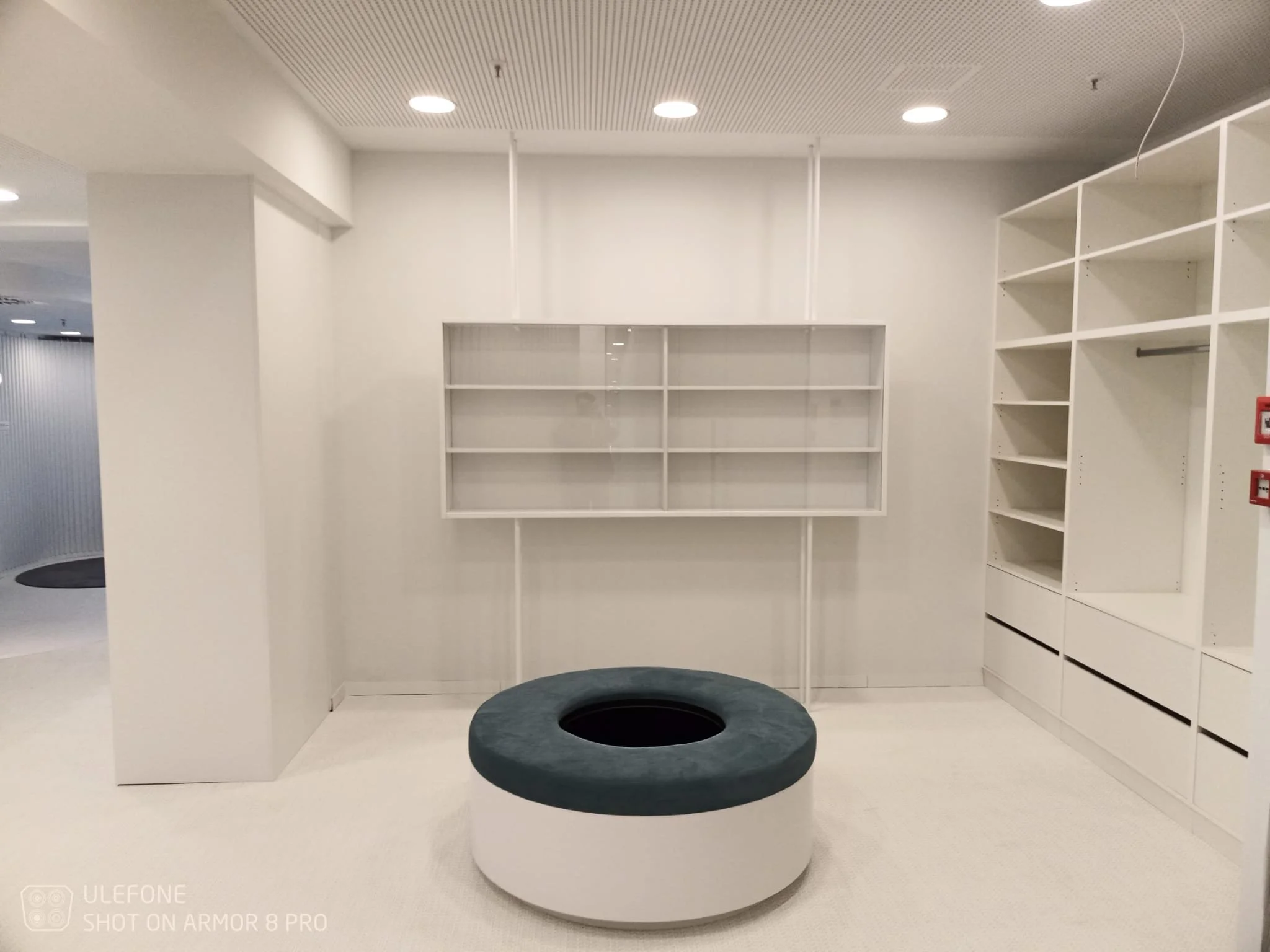 Empty white room with built-in shelves on walls and a cushion-covered round seating in the center.