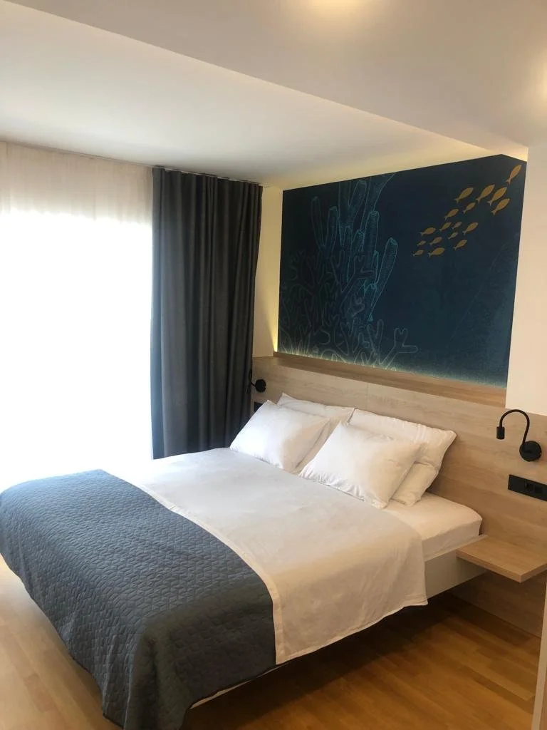 A neatly made bed with white sheets and pillows, topped with a dark gray quilt, in a modern hotel room with a wooden headboard. There is a dark curtain covering a window to the left, and a feature wall with abstract artwork behind the bed. Two black 