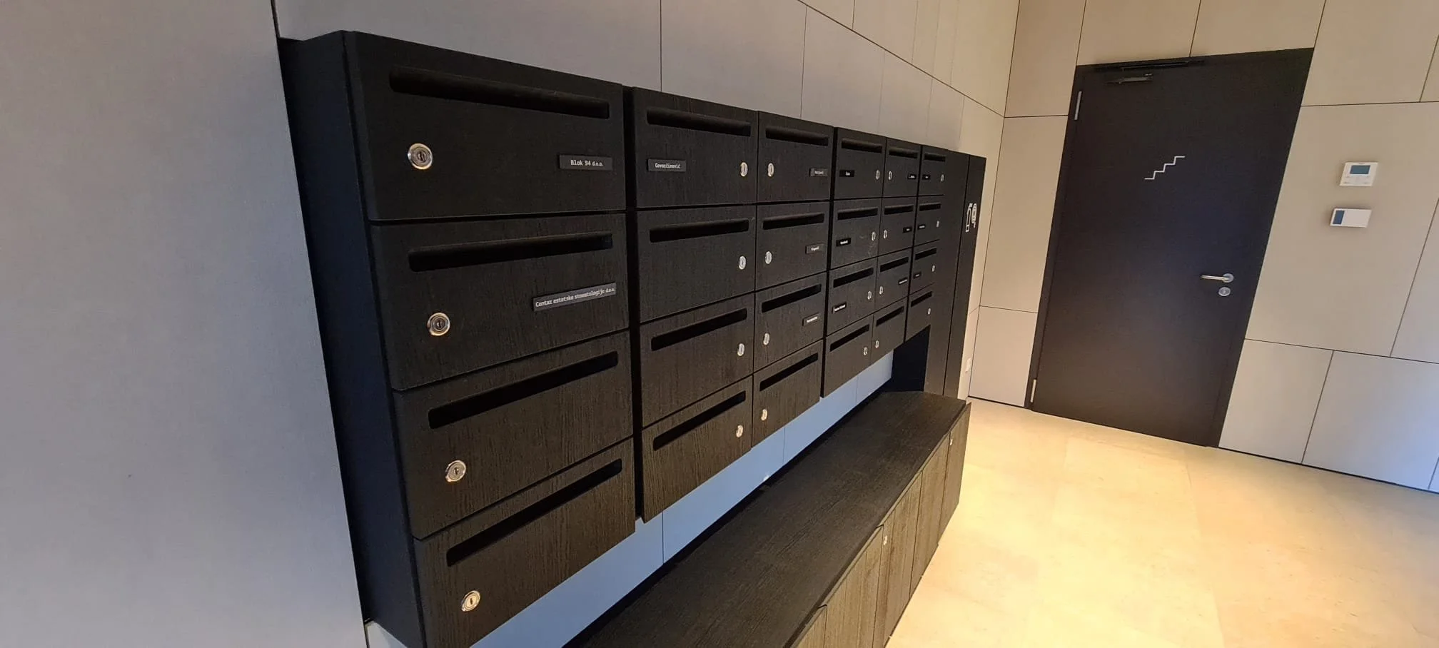 A row of black mailboxes in a modern building hallway with a black door and beige tiled walls and floor.