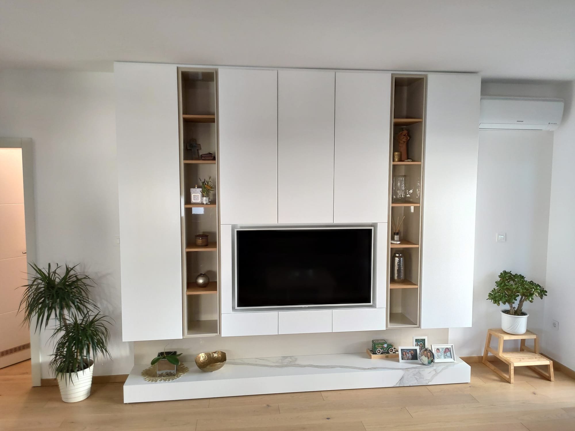 Living room with a white entertainment center, television, potted plants, decorative items, and framed photos.