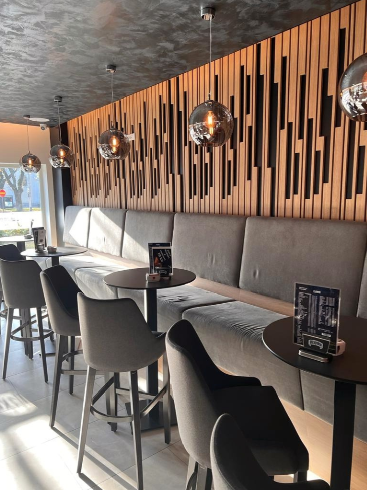 Interior of a modern cafe with high tables, cushioned bench seating, and black chairs. Pendant lights with glass globes hang from the ceiling. A wooden accent wall with vertical slats decorates the space, and sunlight streams through a window.