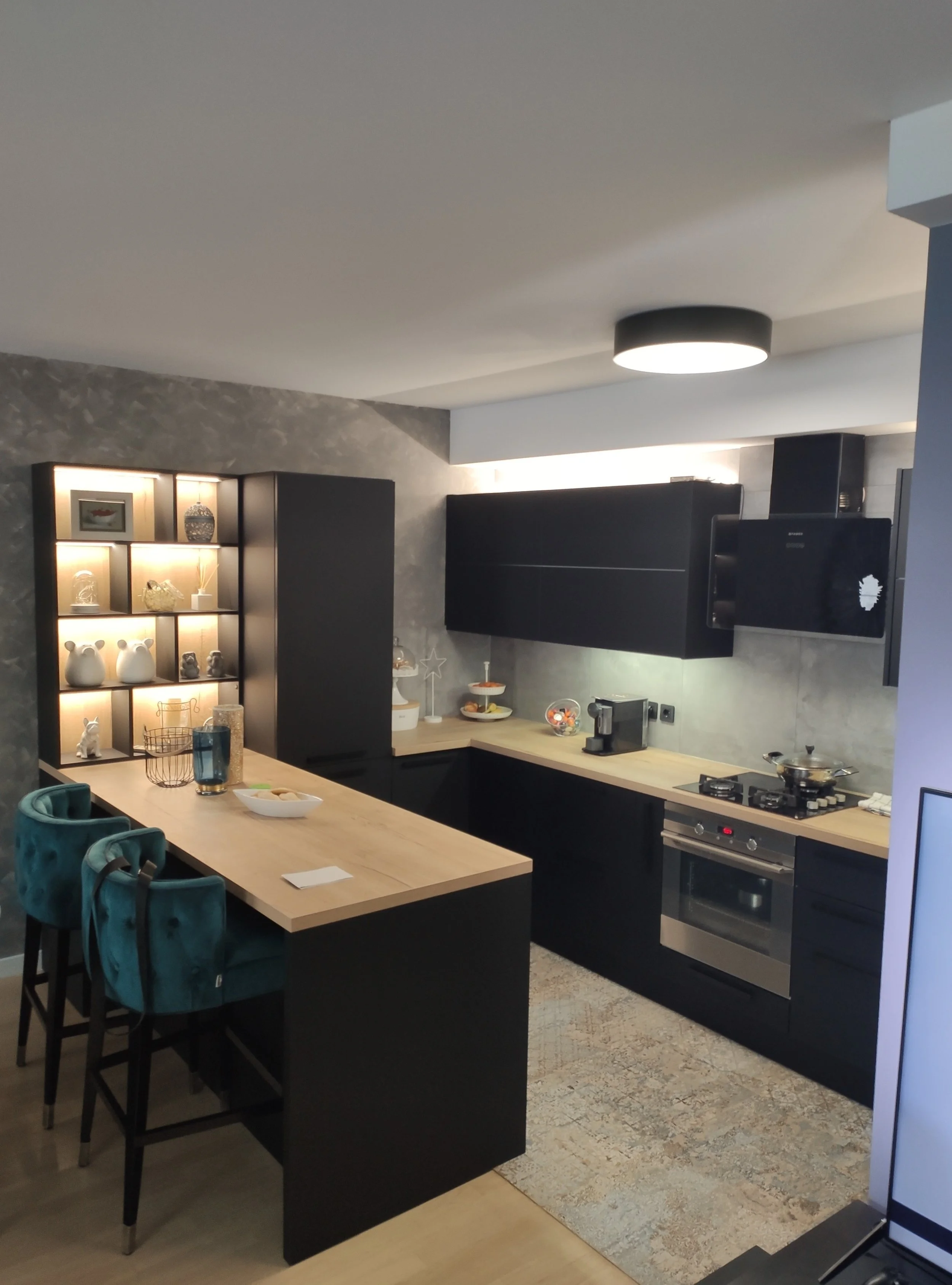 Modern kitchen with black cabinets and light wooden countertops, featuring a small breakfast bar with teal chairs, a tall black storage unit, and decorative items on a shelf.