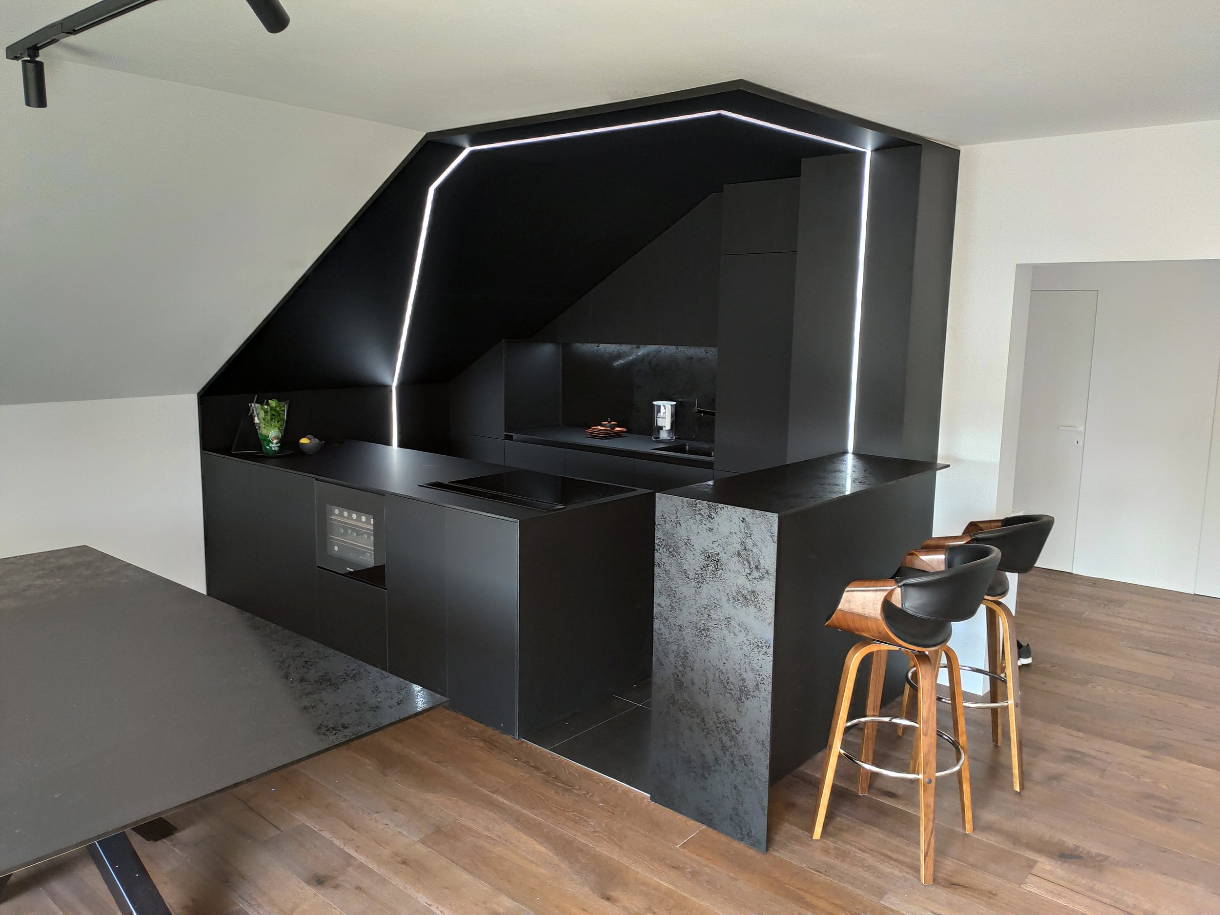 Modern kitchen with black cabinetry and a black upper wall installation with integrated LED lighting, wooden bar stools, and a white door in the background.