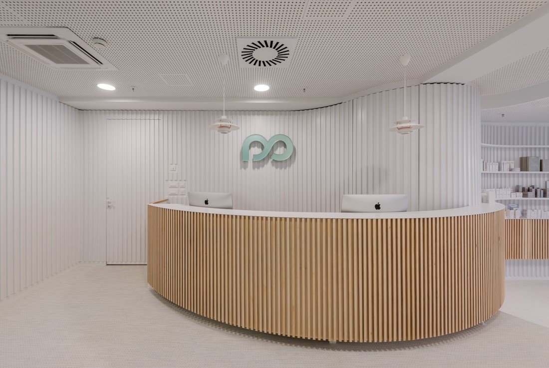 Modern reception area with white walls and ceiling, two white iMac computers on a curved wooden reception desk, and a minimalist logo on the wall behind the desk.