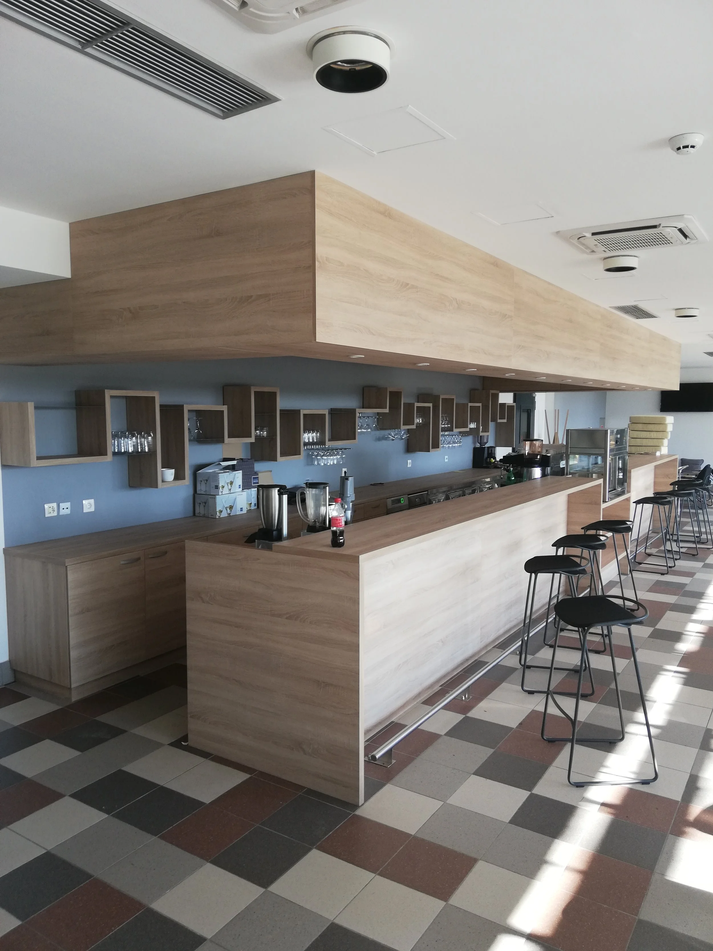 Empty cafe or bar with a long light wood counter, black bar stools, and gray wall with wood shelf decor.
