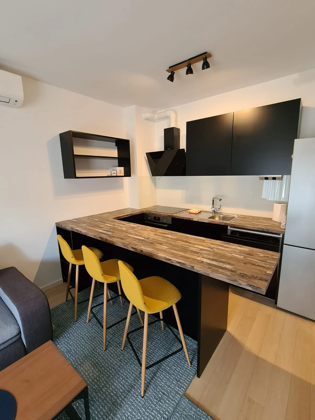 Modern kitchen with black cabinets, wooden countertop, yellow barstools, and appliances including a refrigerator, oven, and range hood.