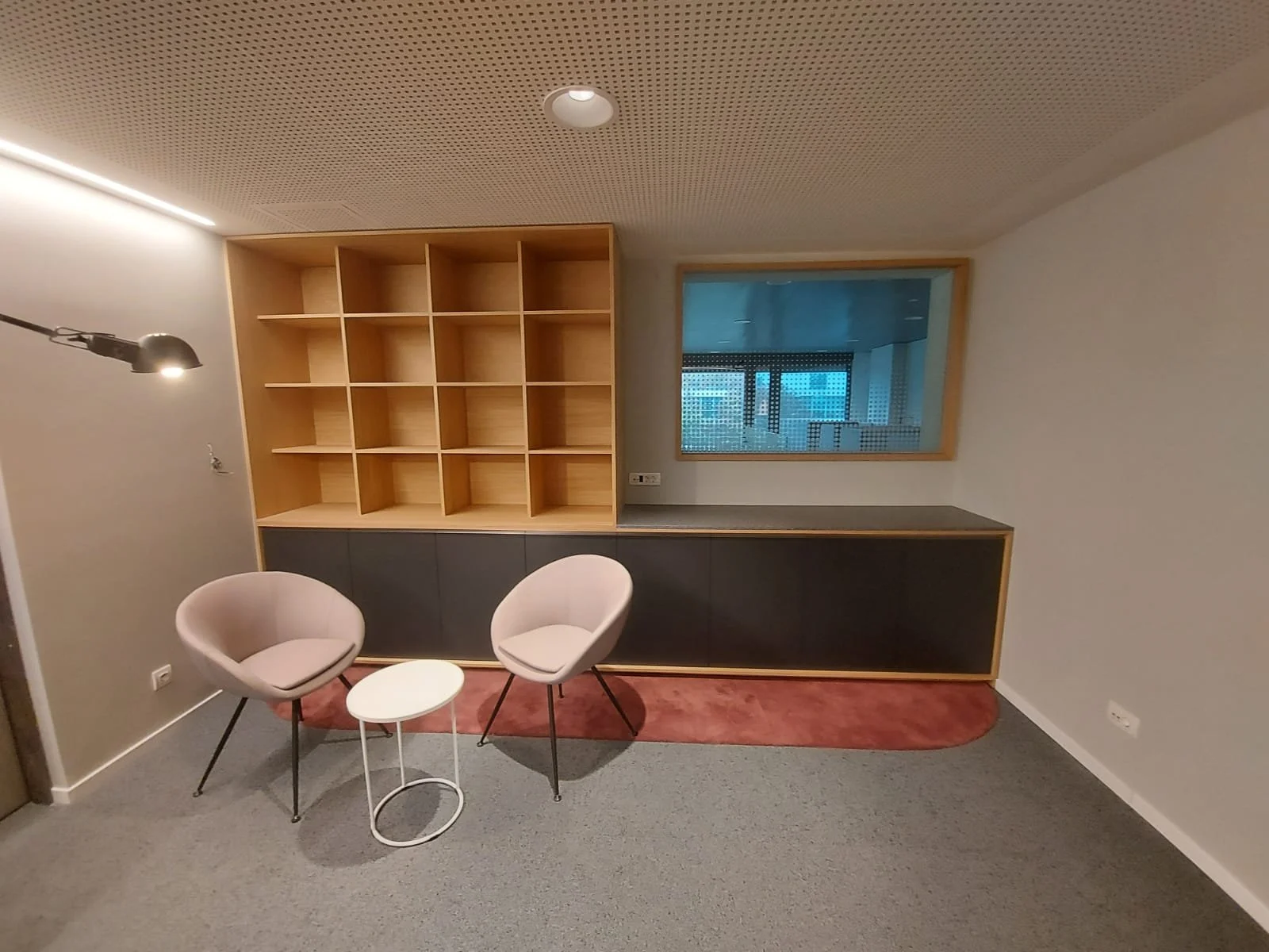 A modern room with two beige armchairs facing each other, a small white round table between them, a built-in wooden bookshelf with open compartments, a window with a view, and a red rug on a gray carpeted floor.