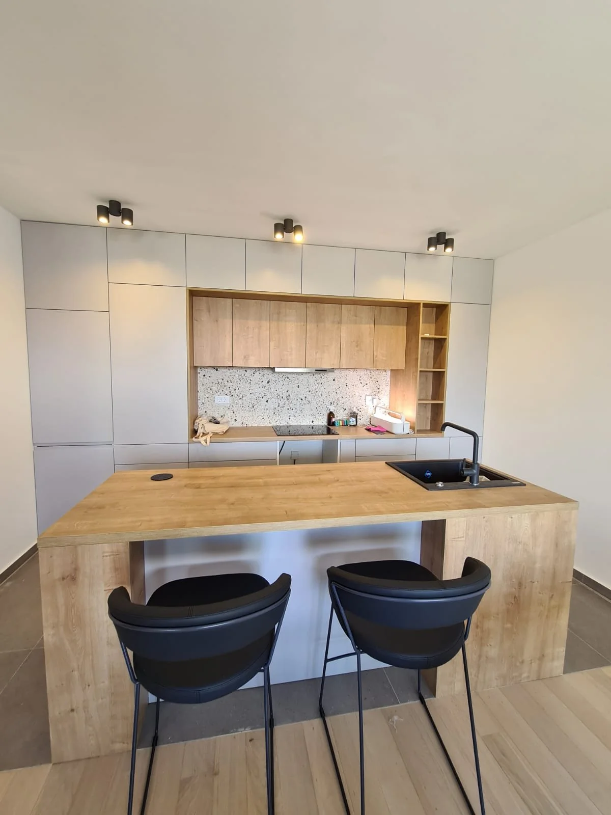 Modern kitchen with wooden island, black chairs, white cabinets, and minimal decor.