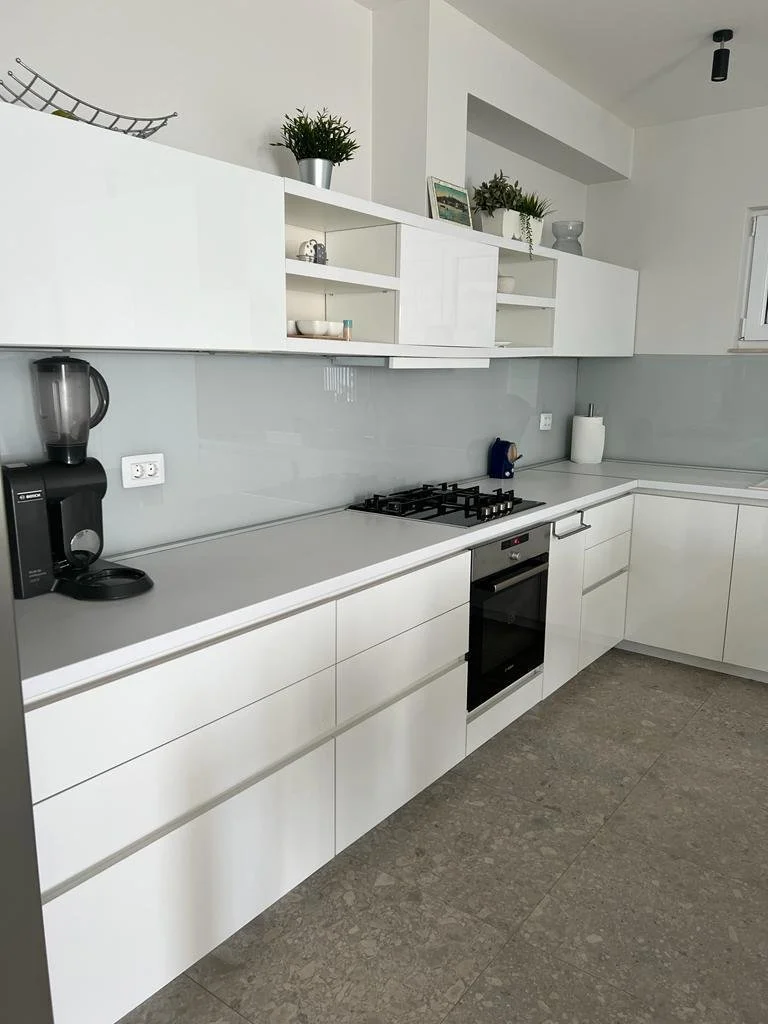 Modern white kitchen with upper and lower cabinets, a built-in oven, and a black coffee maker on the countertop. Open shelves hold dishes and decorative items, and there are plants and framed pictures on top of the upper cabinets.