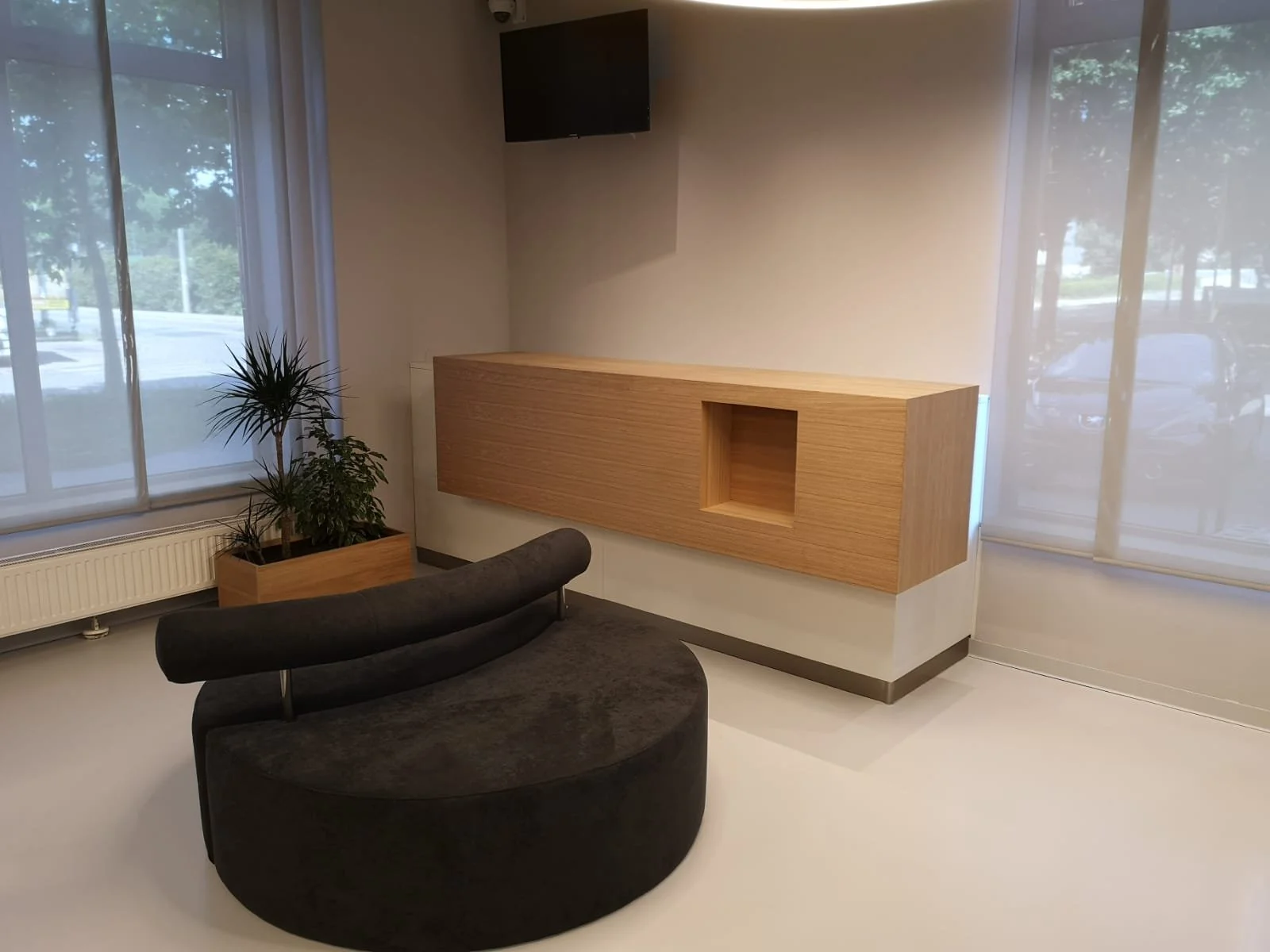 Empty modern waiting area with a black curved chair, a wooden planter with green plants, a wall-mounted wooden cabinet, large windows with blinds, and a wall-mounted flat-screen TV.