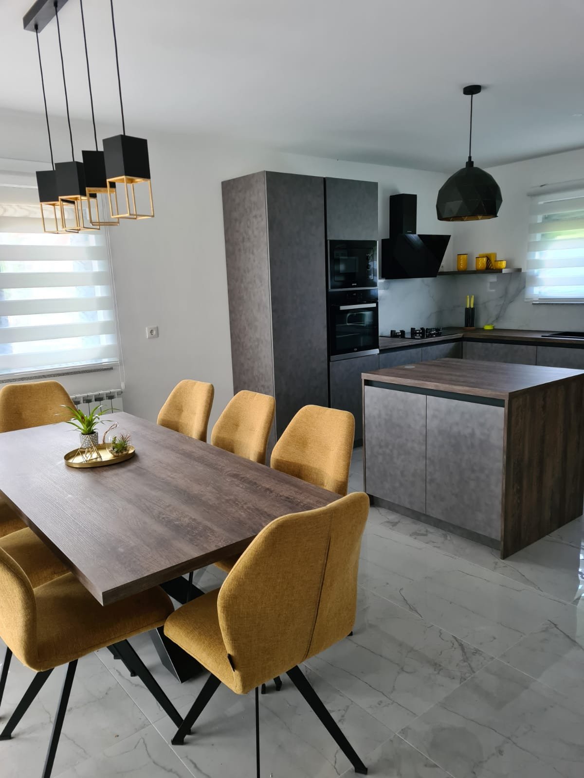 Modern kitchen and dining area with a wooden table, yellow chairs, black hanging pendant lights, gray cabinetry, and marble flooring.
