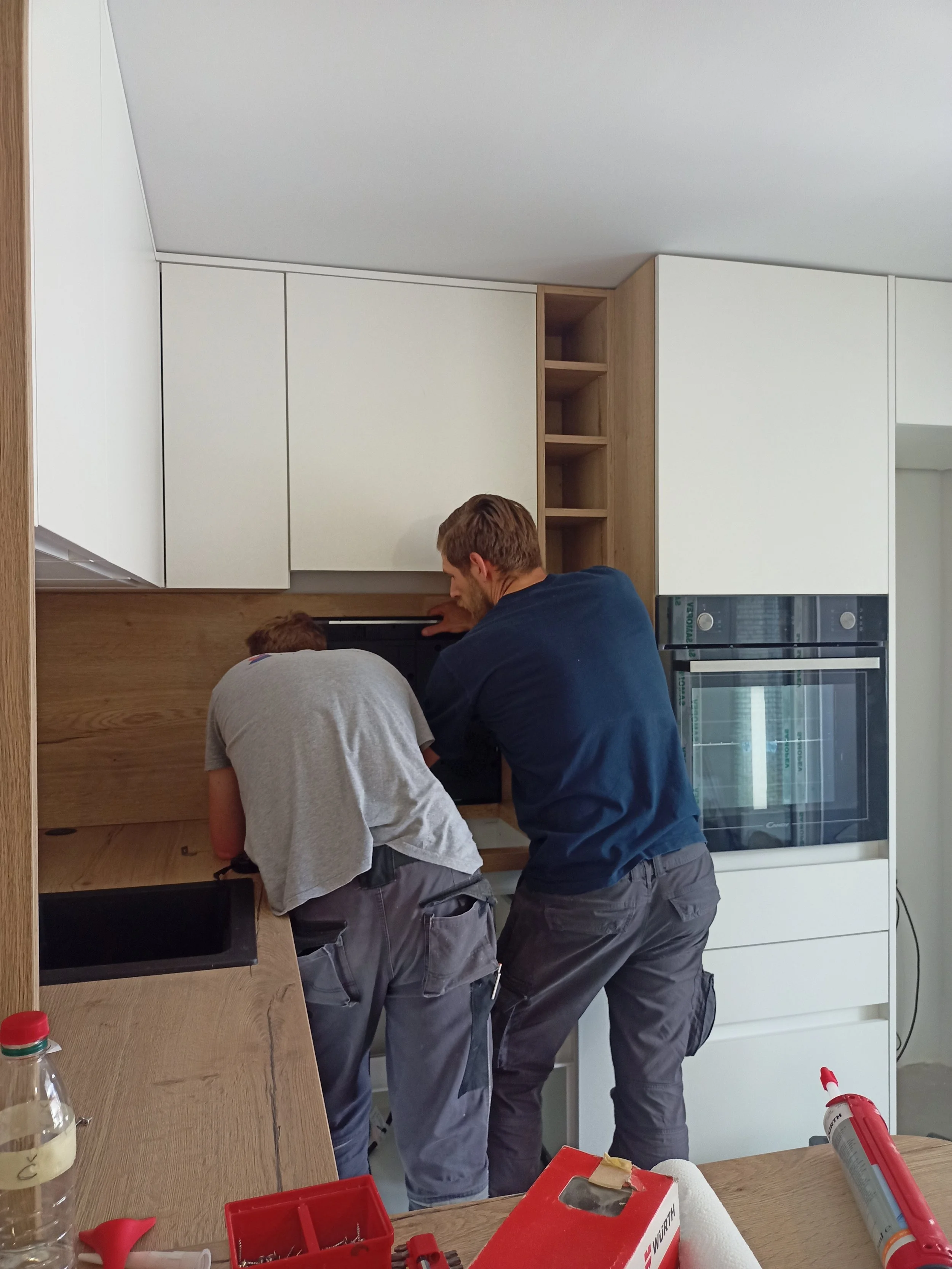 Two men installing or repairing an oven in a modern kitchen with white cabinetry and wooden accents.
