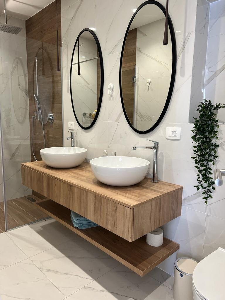 Modern bathroom with a floating wooden vanity, two white vessel sinks, and two large oval mirrors. There is a shower area with a glass door, wall-mounted shower heads, and a wooden accent wall. A small plant is hanging on the wall, and a white toilet