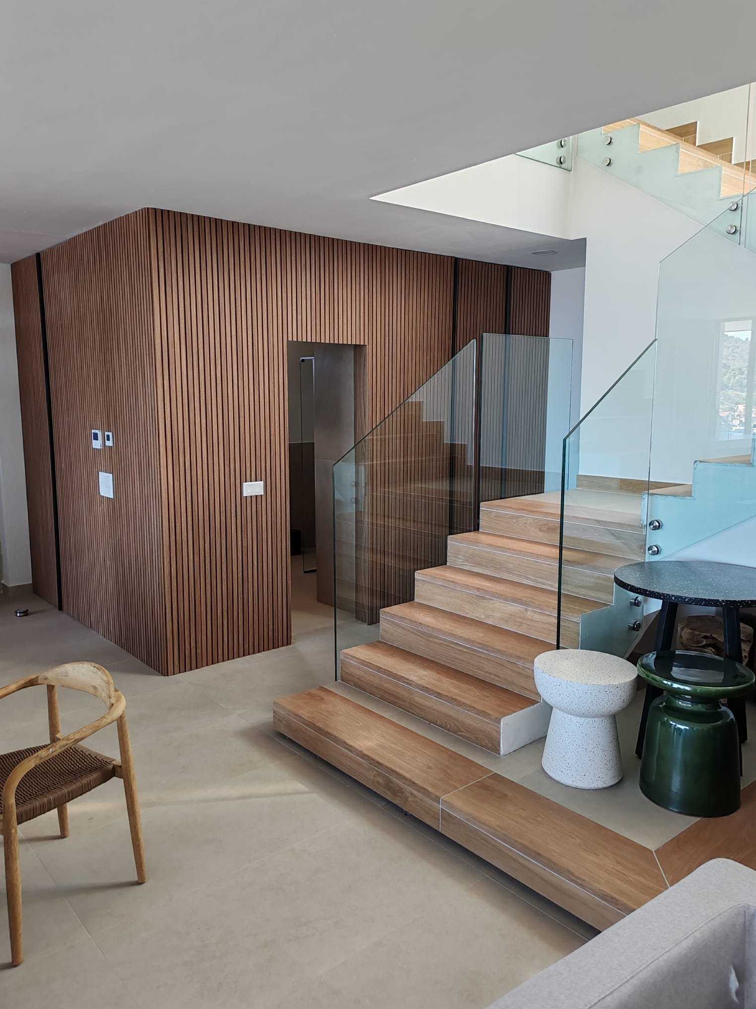 Interior view of a modern home featuring a wooden staircase with glass railing, a wood-paneled wall, and minimal decor including a round black table, a green stool, and textured stools.