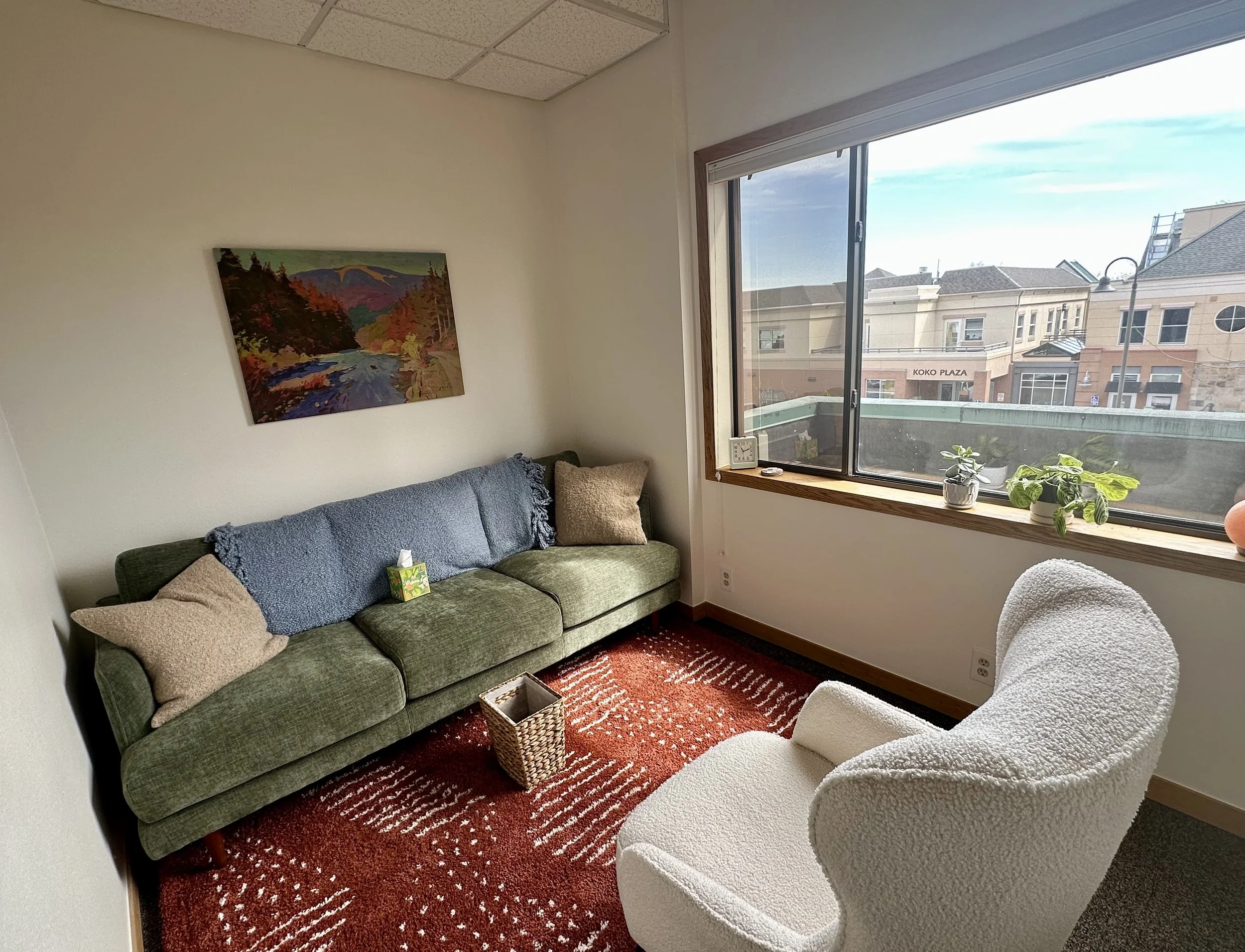 A cozy living room with a green sofa, beige pillows, a white plush armchair, a red patterned rug, and a window showing a cityscape with buildings and a streetlamp.